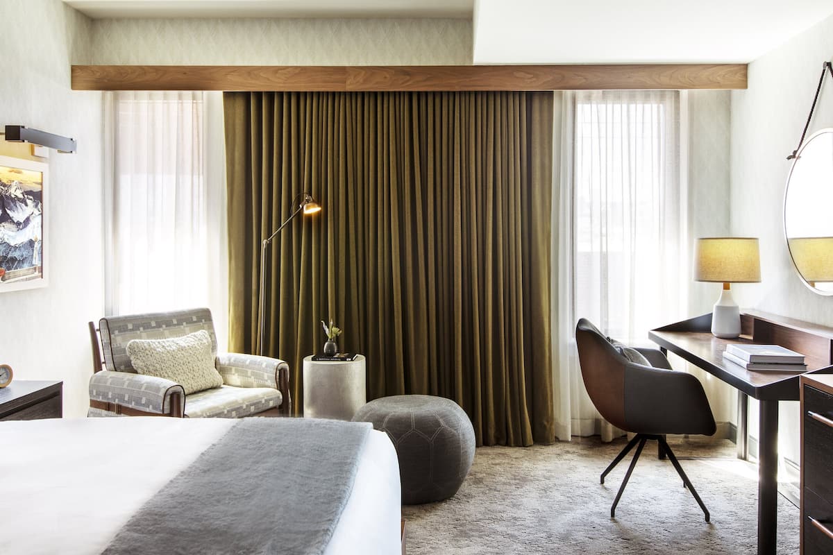 A hotel room with a bed, a desk, a chair, a lamp, and a large window with curtains.