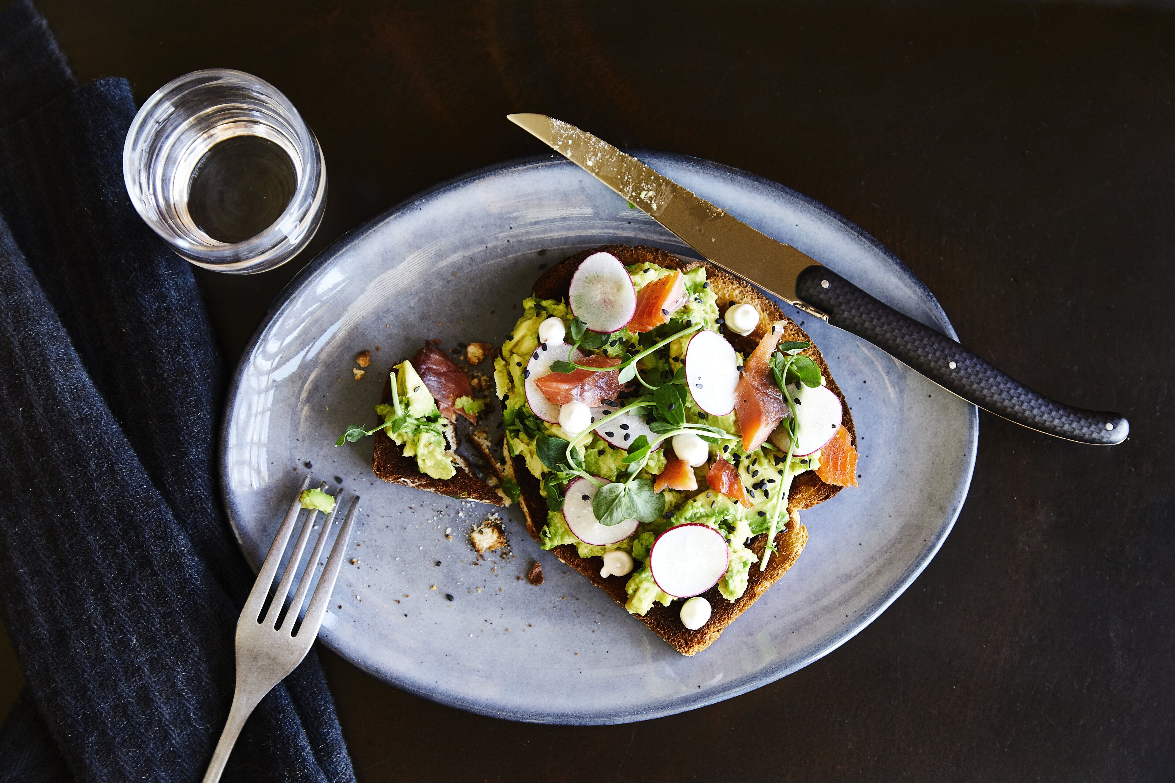 Ein Teller mit Toast, belegt mit Avocado, Rettich und Räucherlachs, dazu Messer und Gabel auf einem dunklen Tisch.