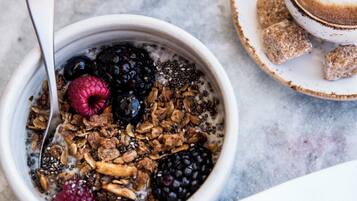 A bowl of cereal with fruits and granola, a glass of orange juice, and a cup of coffee with milk on a marble surface.