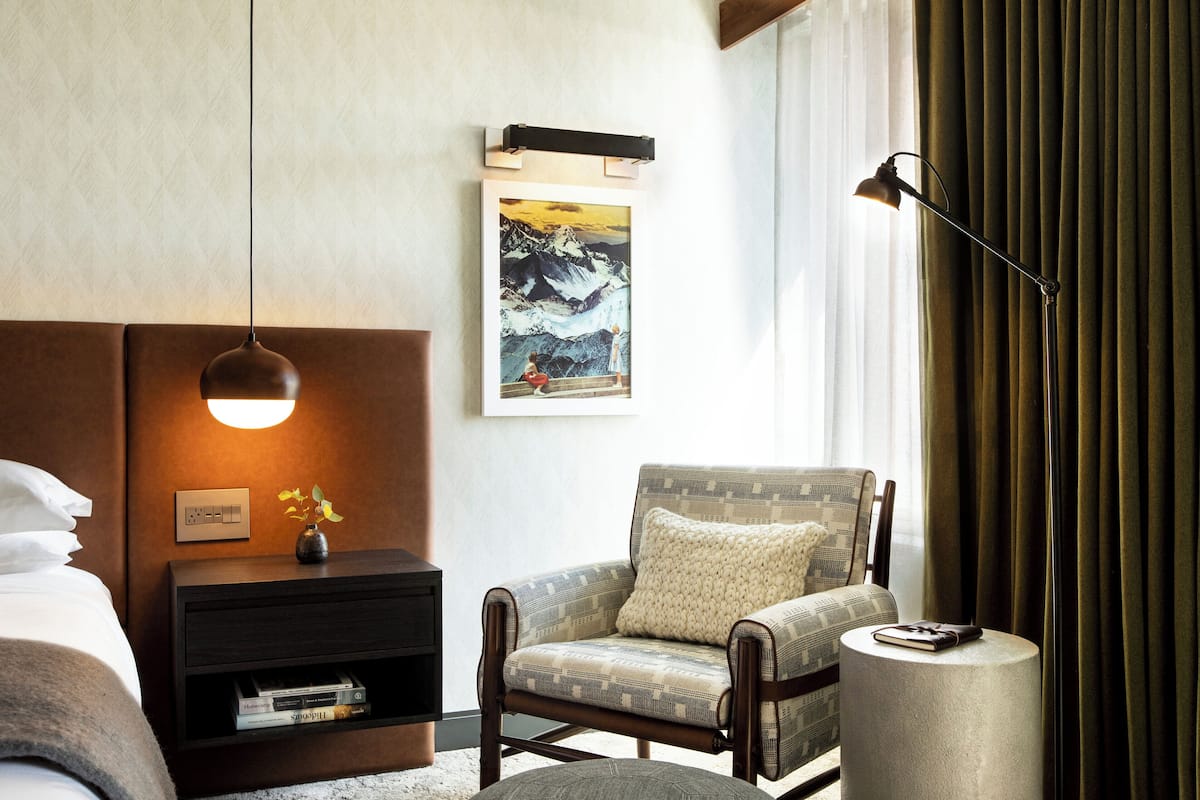 A modern hotel room with a large bed, a nightstand with a lamp, a patterned armchair, and a framed picture on the wall.