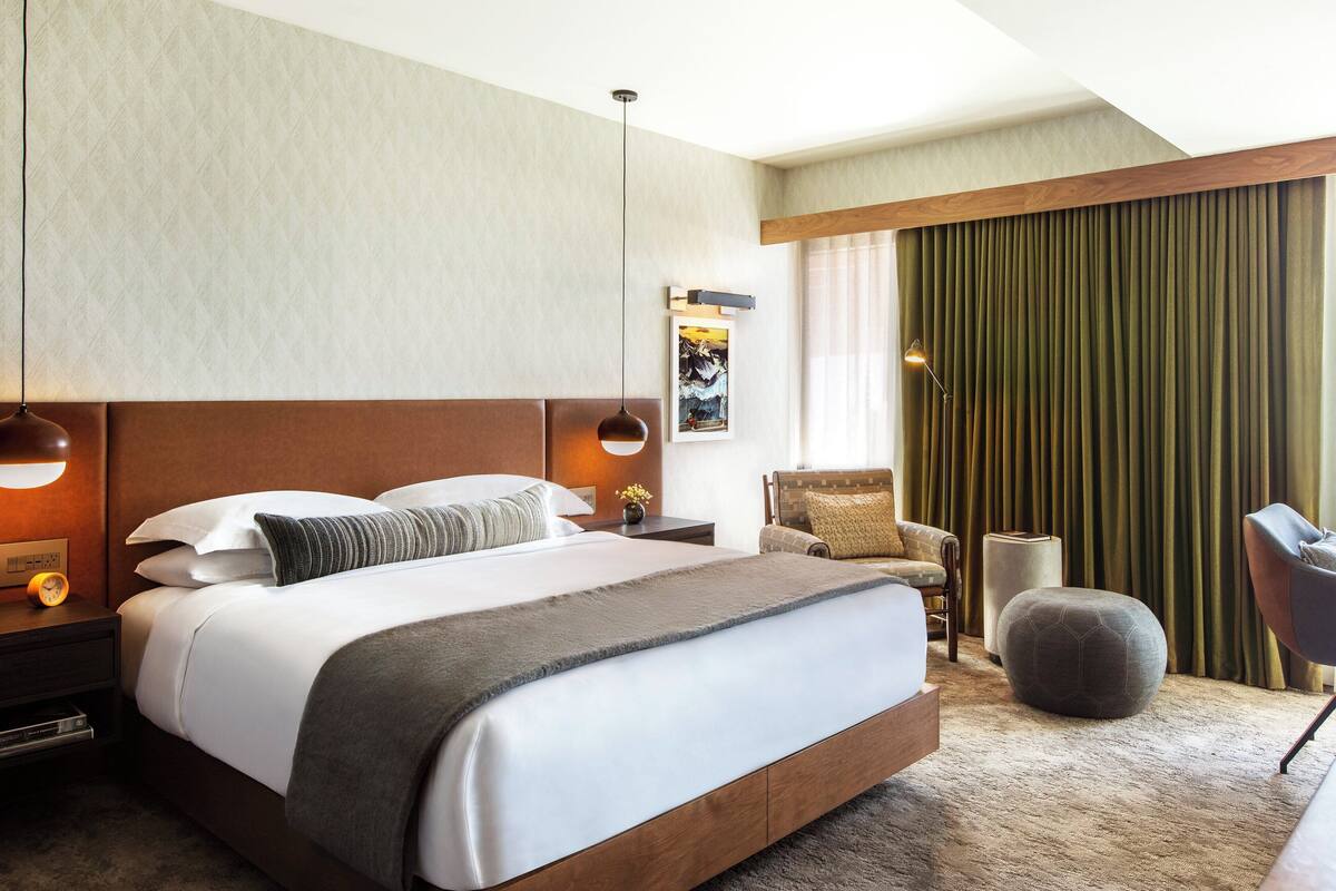 A modern hotel room with a large bed, two bedside tables, a chair, a small ottoman, and a view of the city through a window with curtains.