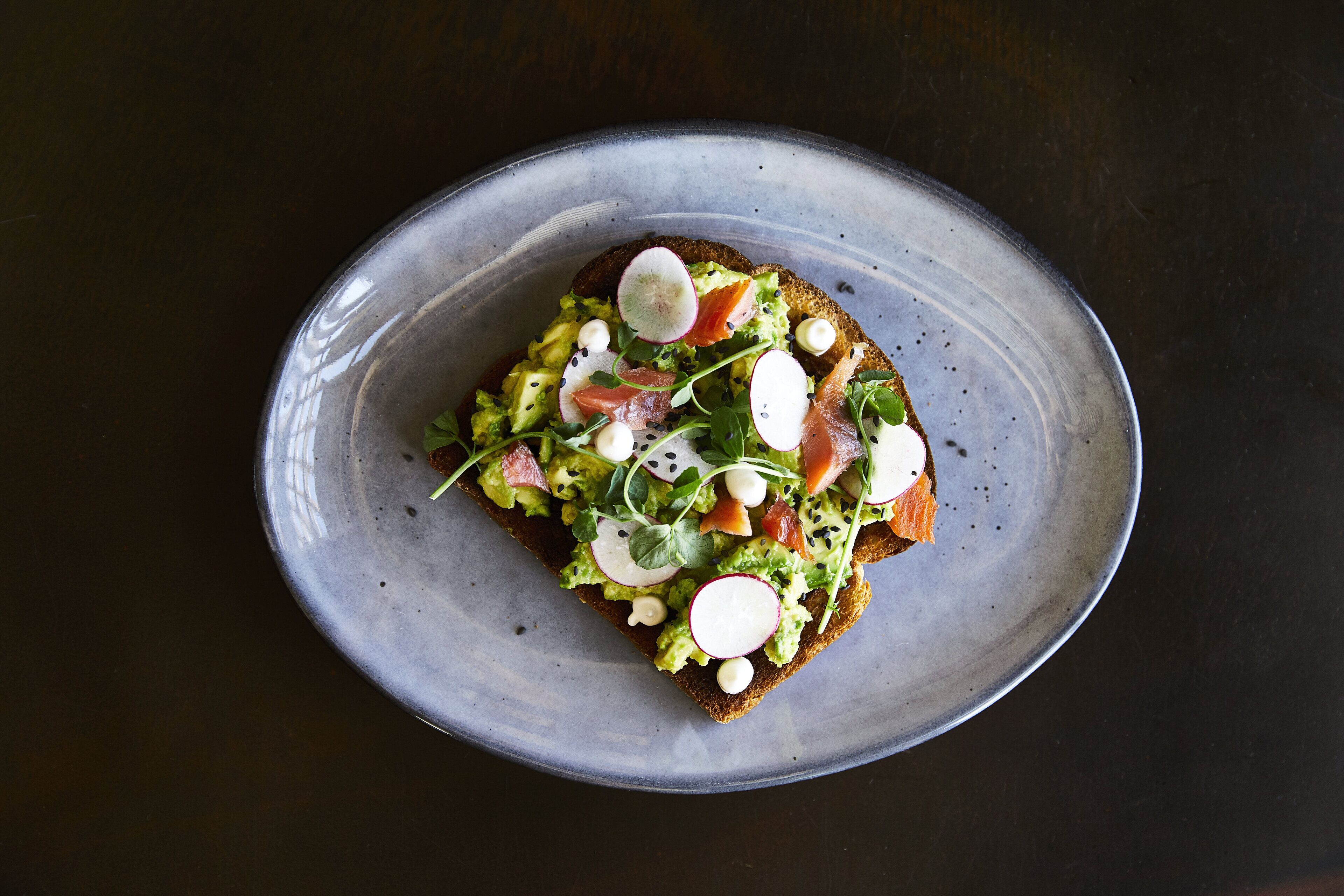 Eine Scheibe Toast mit Avocado, Radieschen und Grünzeug auf einem Keramikteller.
