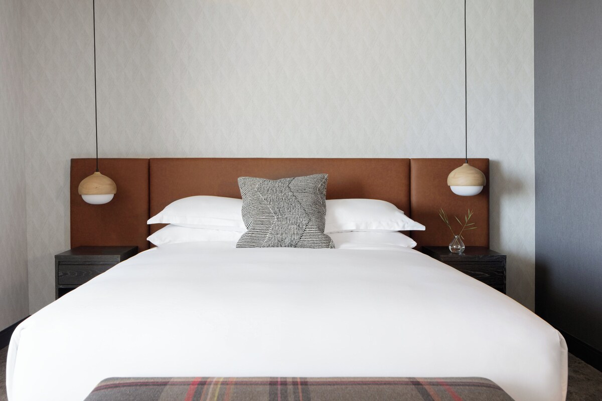 Een netjes opgemaakt bed met een geruit plaid, twee nachtkastjes met lampen, en een effen muur met subtiele textuur.