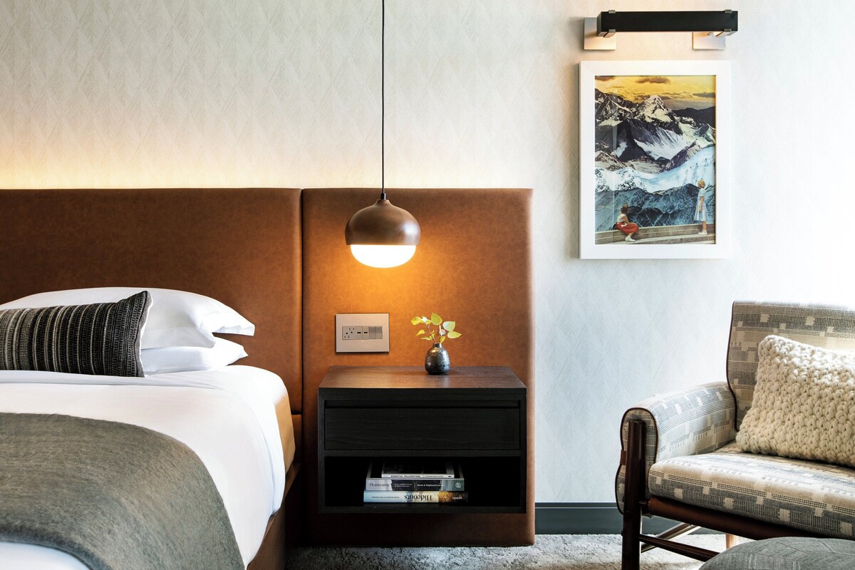 Een hotelkamer met een bed, nachtkastje, lamp, stoel en een ingelijst schilderij aan de muur.