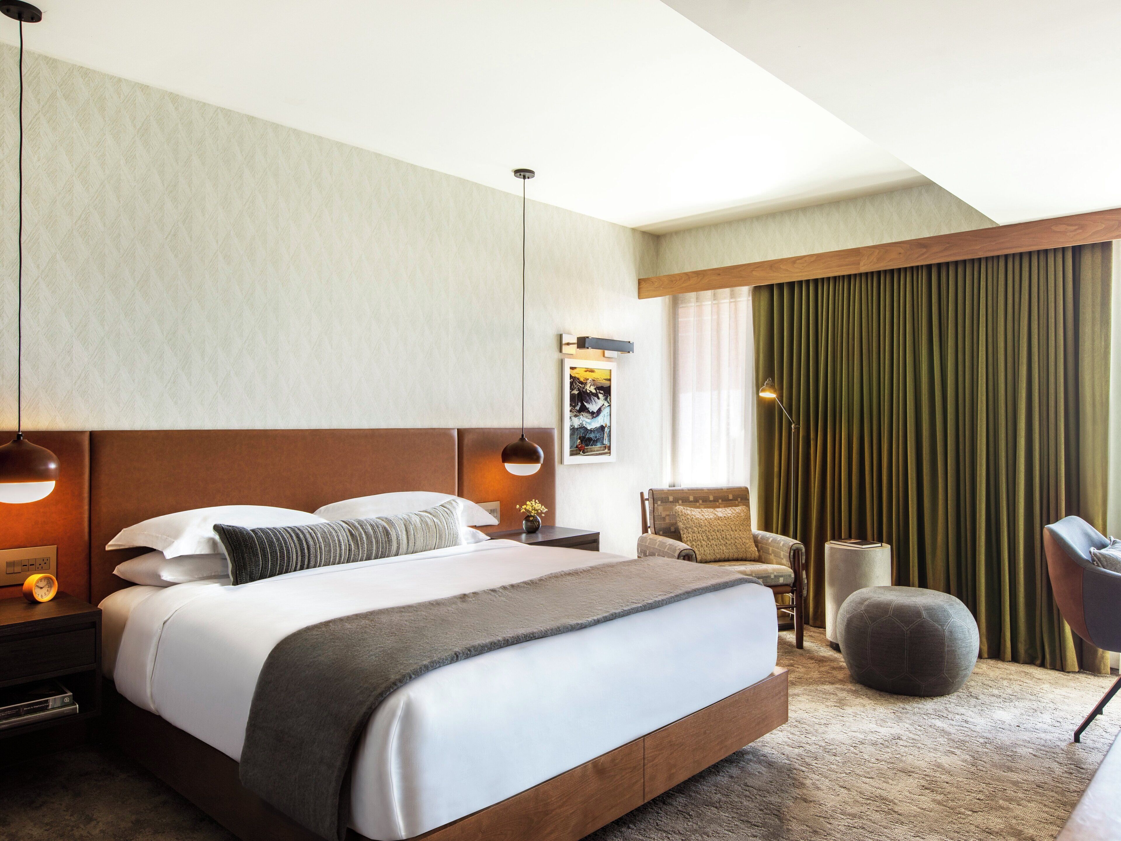 Une chambre d’hôtel moderne avec un grand lit, deux tables de chevet, une chaise, un petit pouf et une vue sur la ville par une fenêtre agrémentée de rideaux.