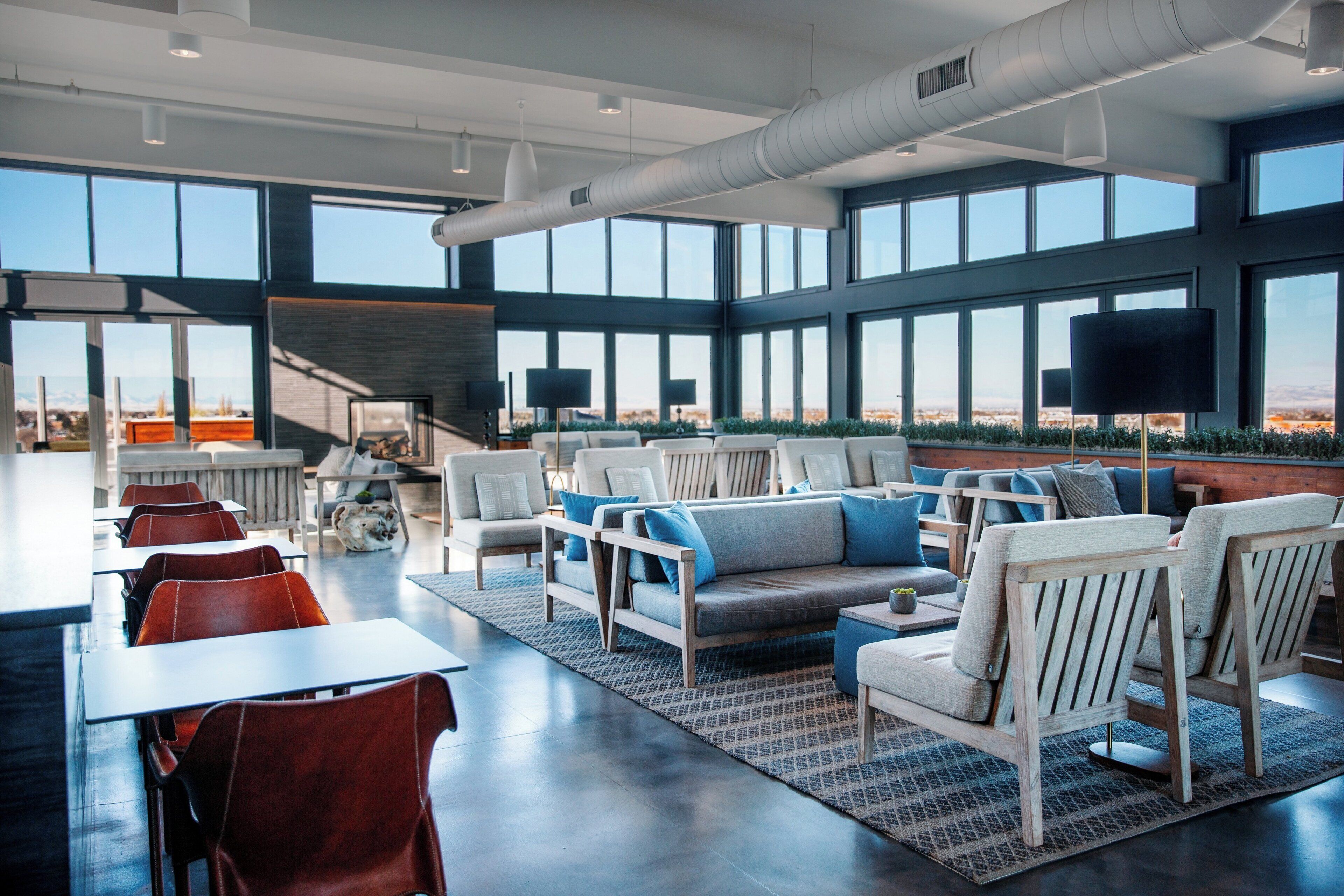 A modern lounge area with seating and tables, large windows offering a view, and a staircase leading to an upper level.