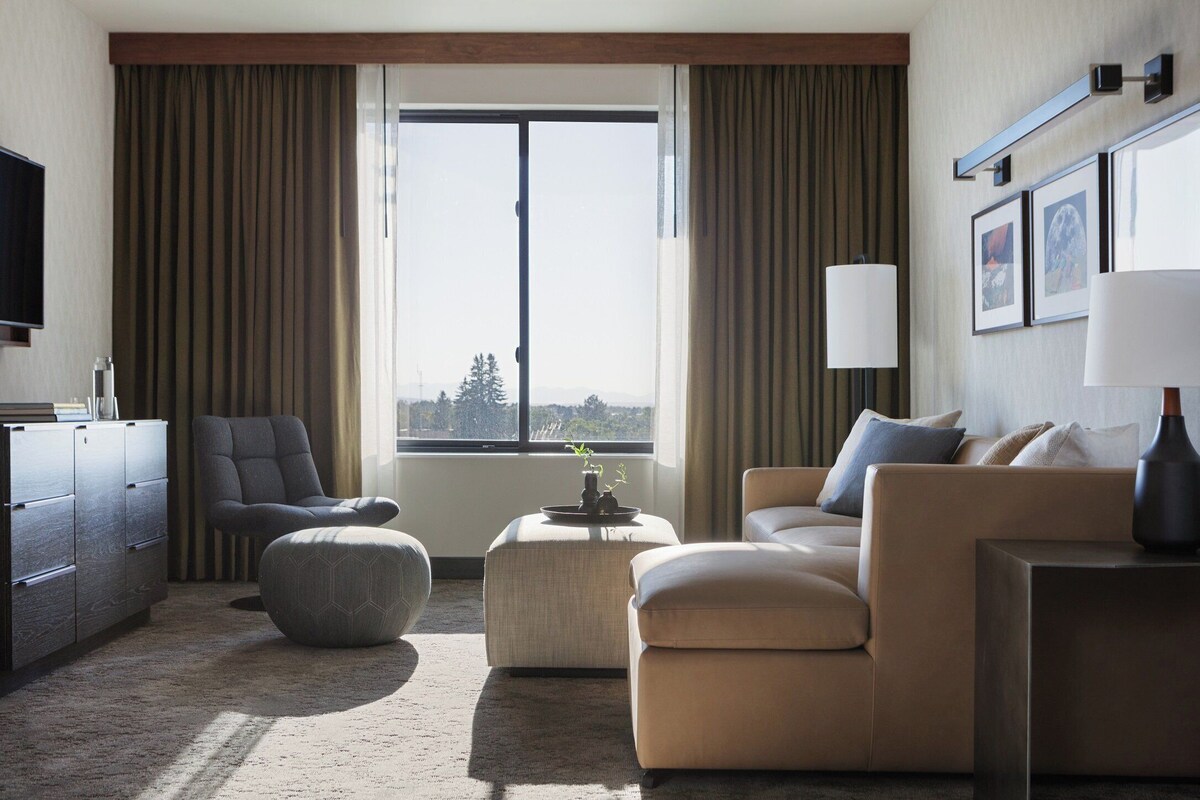 Een moderne hotelkamer met een groot raam, een bank, een stoel, een klein voetenbankje en een televisie op een kast.
