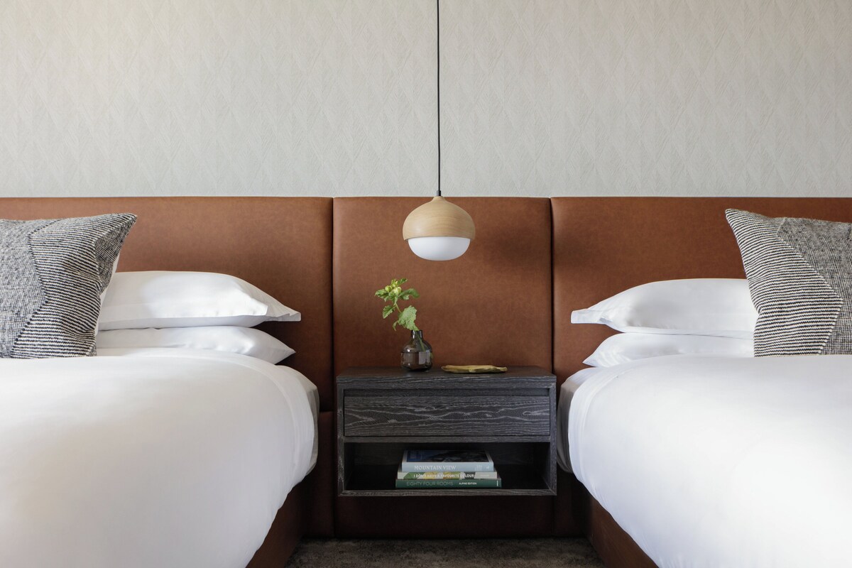 Een hotelkamer met twee bedden, een nachtkastje met een vaas en boeken, en een hanglamp.