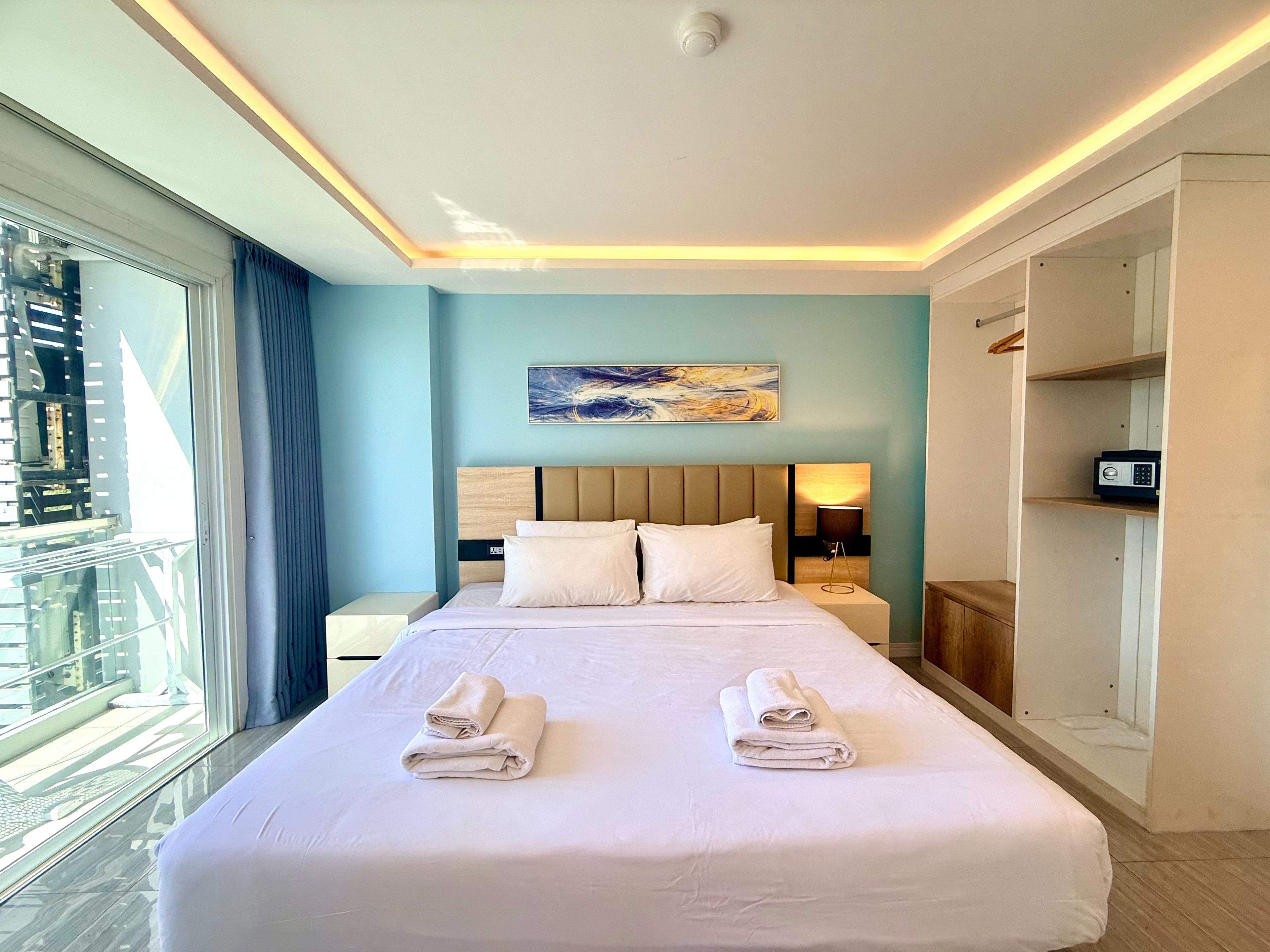 A hotel room with a large bed, a bedside table, a lamp, a painting, and a view of the outside.