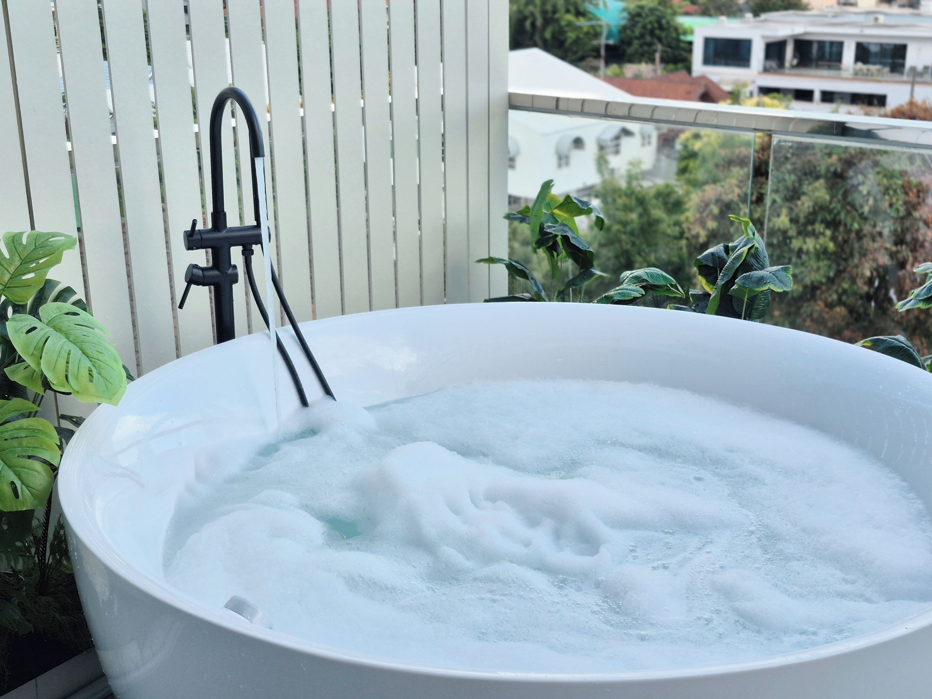 A hot tub with a faucet, surrounded by plants, on a balcony with a view of buildings.