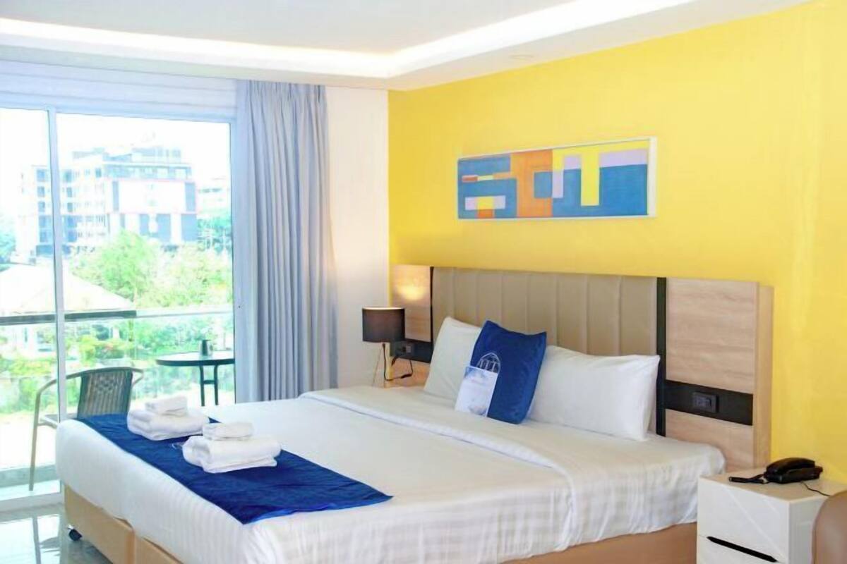 A hotel room with a bed, a bedside table, a lamp, a phone, and a view of the outdoors.