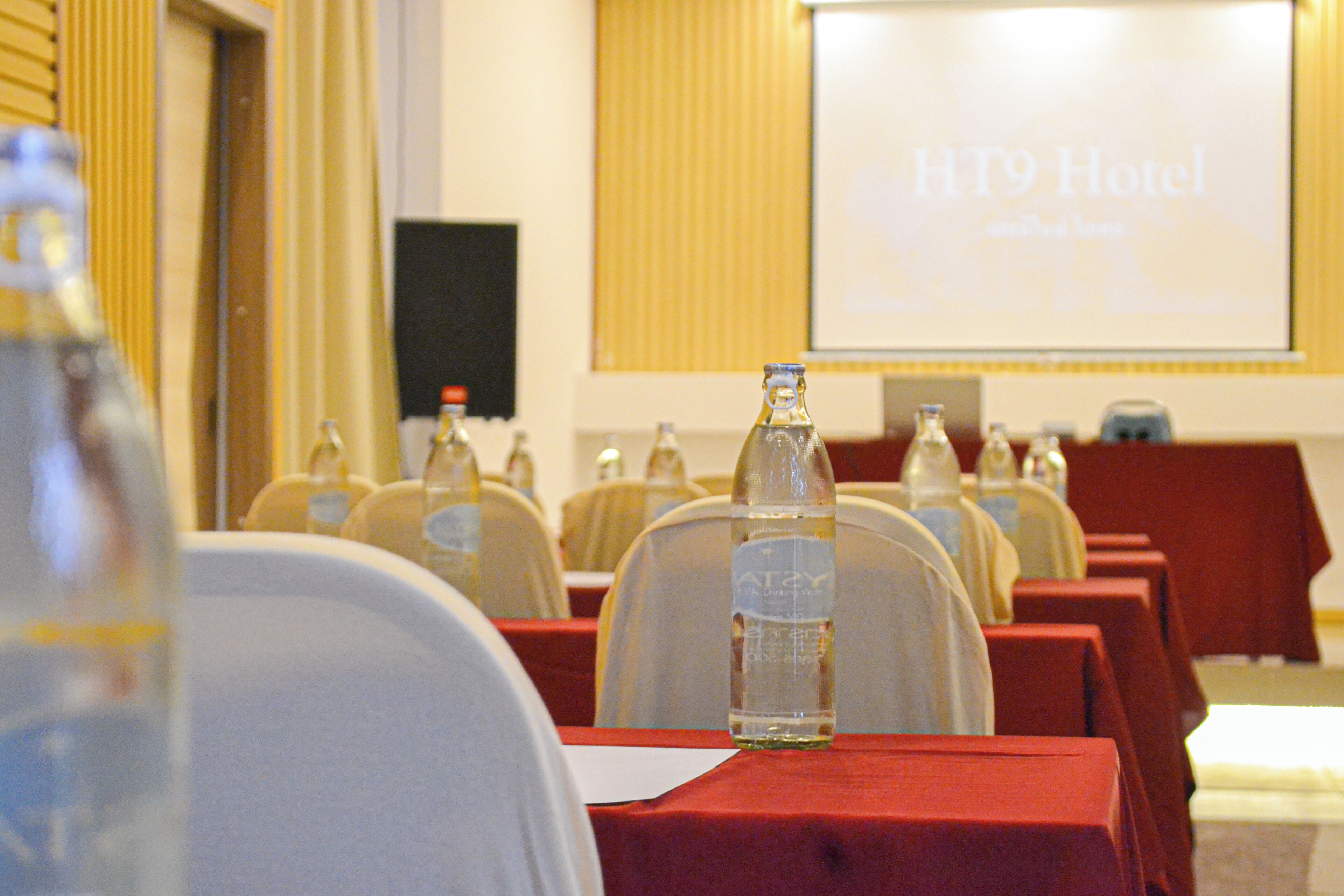 A conference room with rows of tables, each with a bottle of water and a red tablecloth. A projector screen in the background displays "HT9 Hotel" with a tagline.
