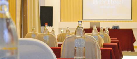 A conference room with rows of tables, each with a bottle of water and a red tablecloth. A projector screen in the background displays "HT9 Hotel" with a tagline.