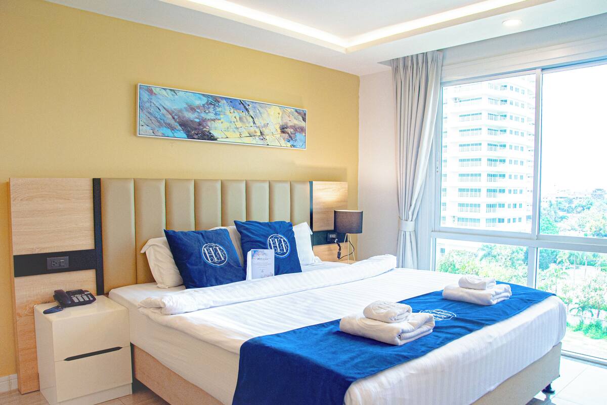 A hotel room with a large bed, a nightstand with a phone, a painting on the wall, and a window with a view of buildings.