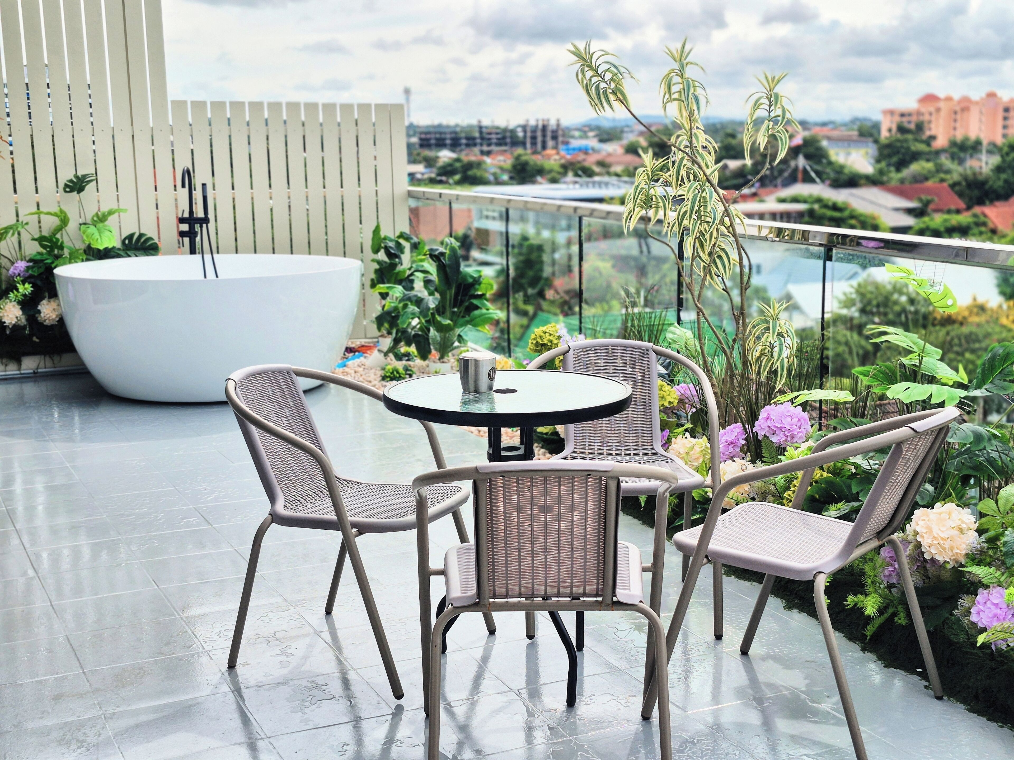 A rooftop terrace with a round table and four wicker chairs, a white bathtub, and various potted plants.