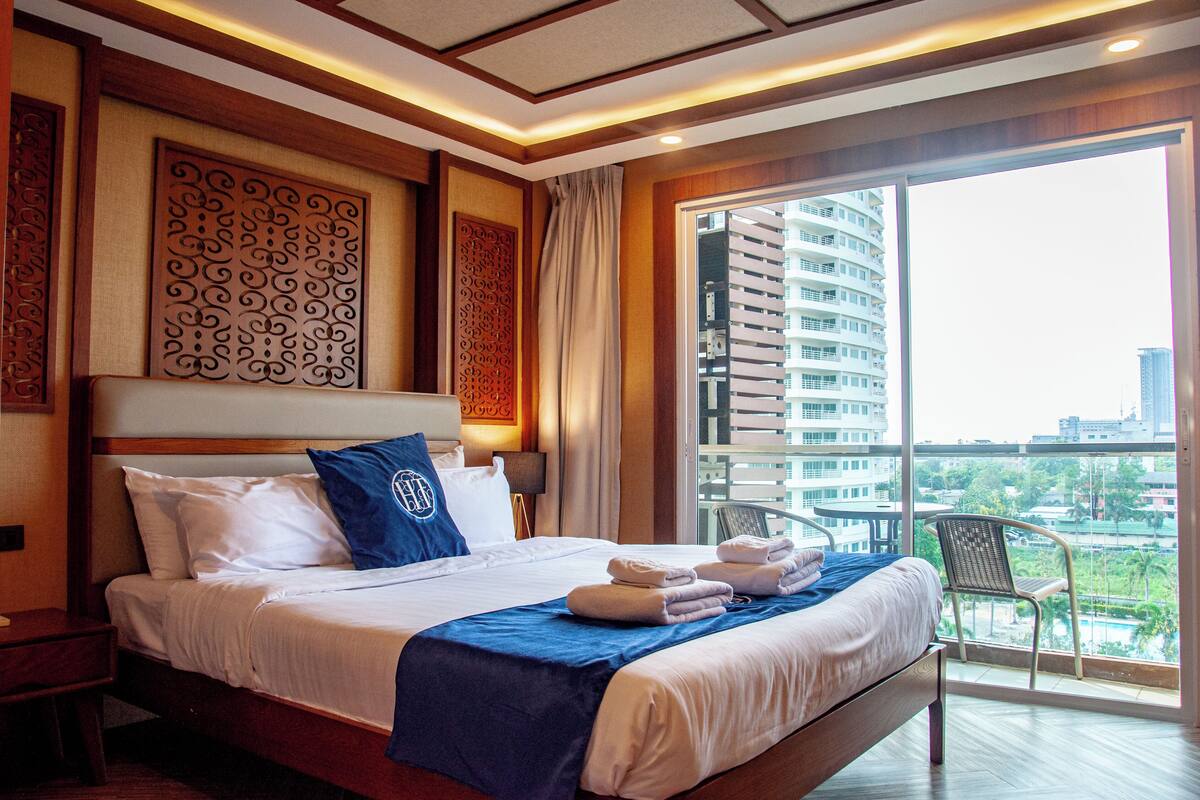 A hotel room with a large bed, a view of a cityscape through a balcony, and a decorative wall panel.