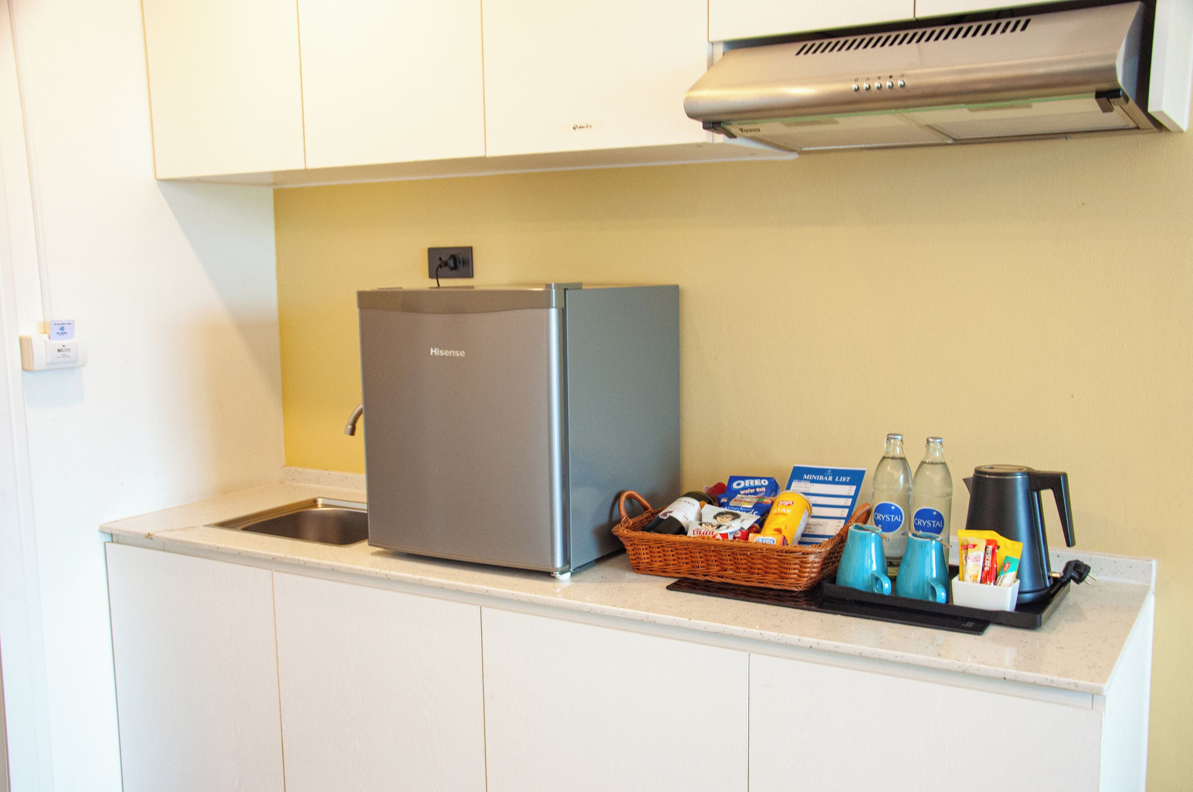 A compact kitchen with a stainless steel refrigerator, a sink, and a countertop with a basket containing food items and a coffee maker.