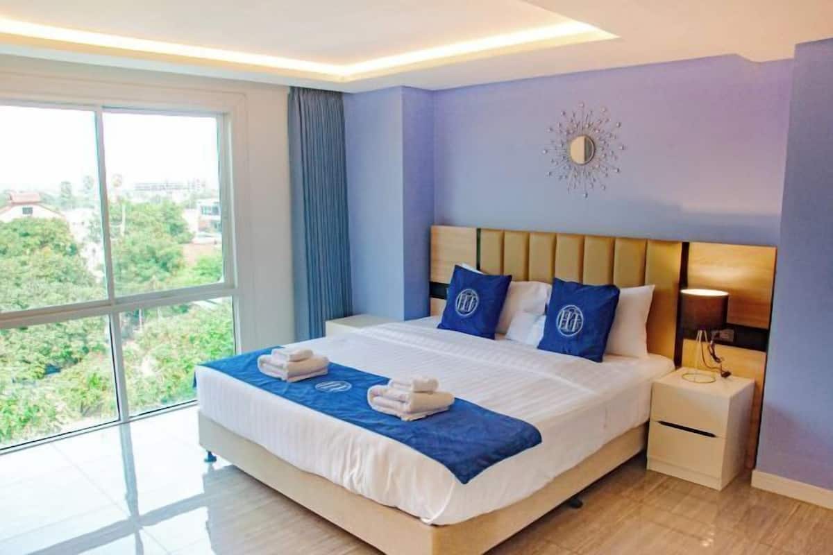 A hotel room with a large bed, a nightstand, a wall-mounted mirror, and a view of greenery outside.