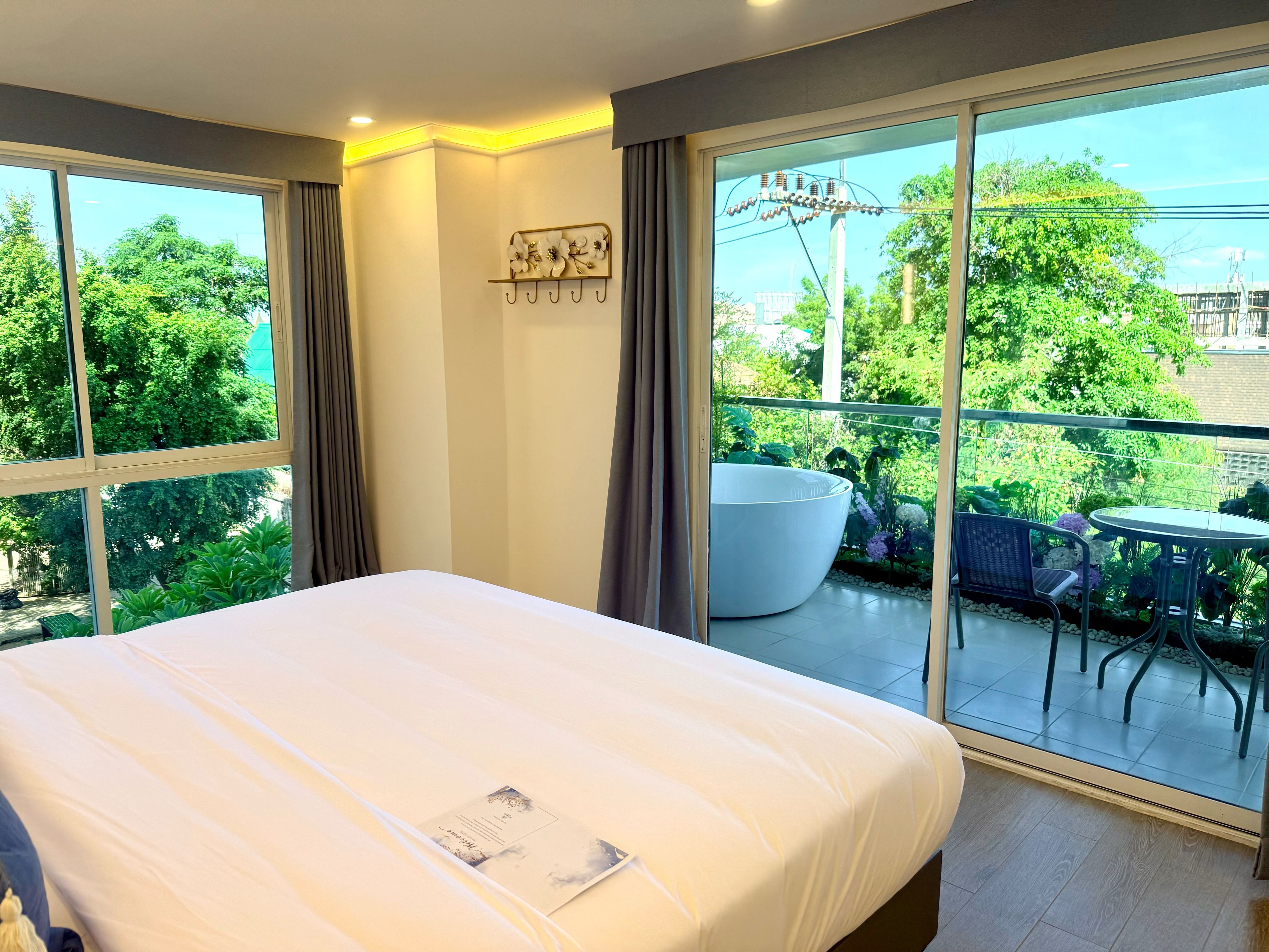A hotel room with a large bed, a balcony with a table and chairs, and a view of greenery and buildings.