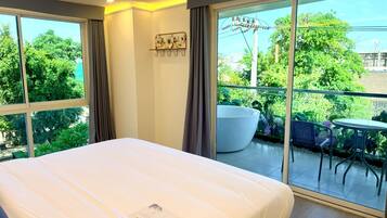 A hotel room with a large bed, a balcony with a table and chairs, and a view of greenery and buildings.