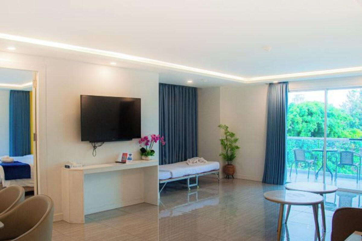 A modern hotel room with a flat-screen TV, a bedside table with a plant, a bed, a small desk, and a balcony with outdoor seating.