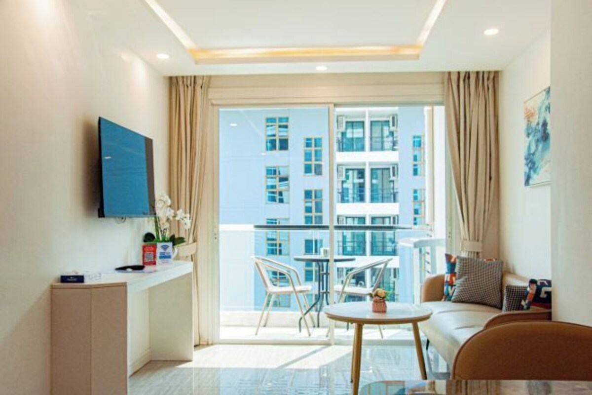 A modern living room with a flat-screen TV, a sofa, a dining table, and a balcony with a city view.