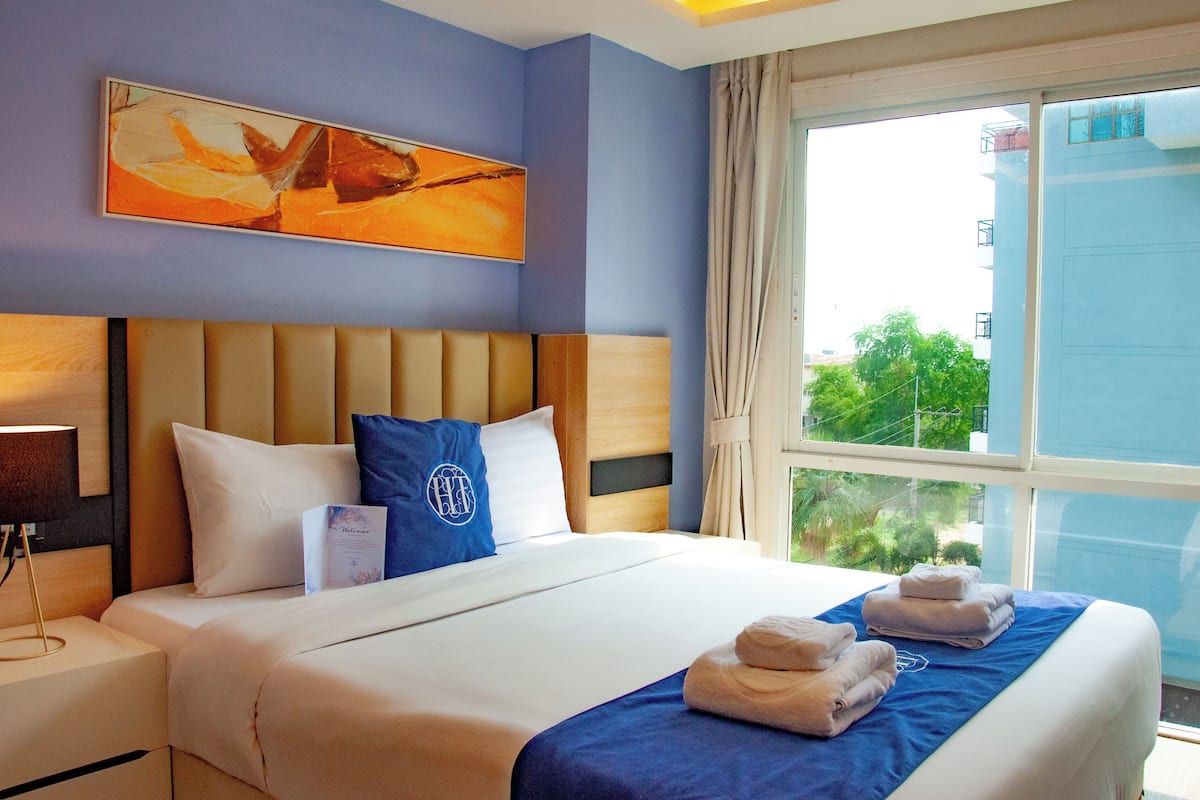 A hotel room with a large bed, a headboard, a window with a view of greenery, and a painting on the wall.