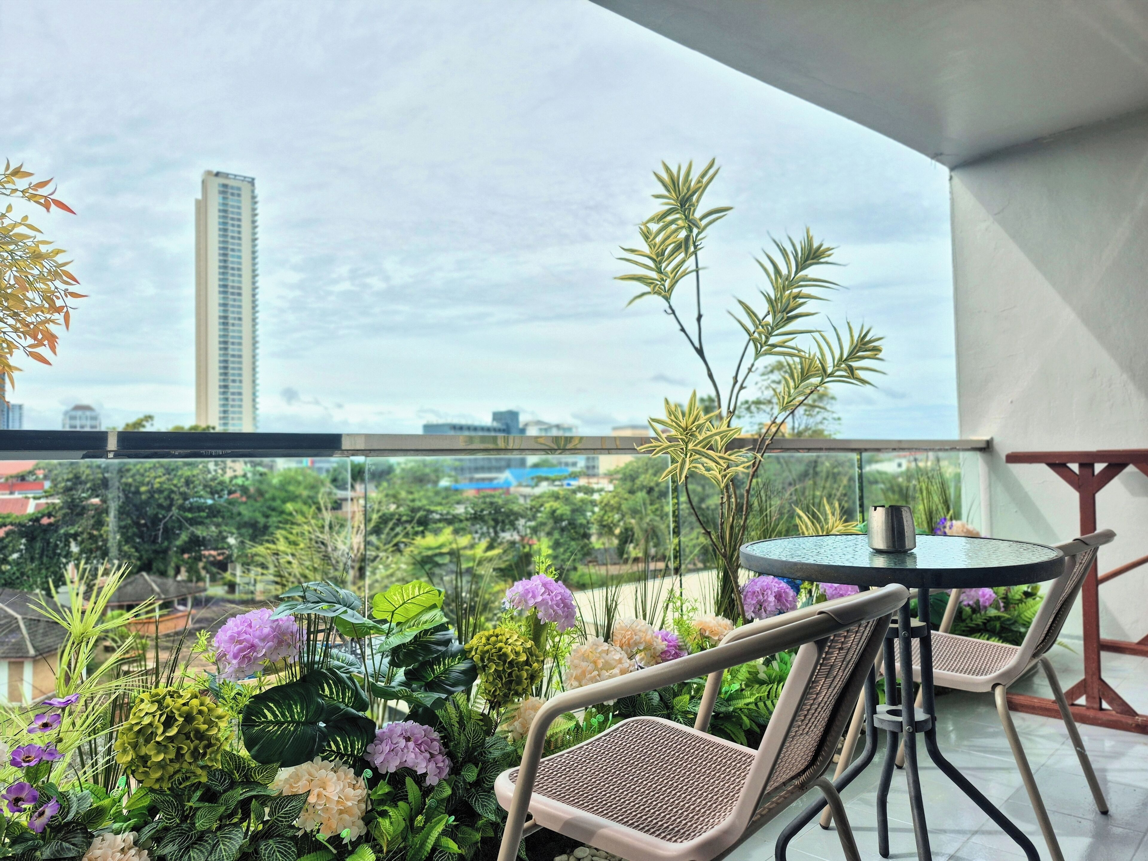 A balcony with a round table and two chairs, surrounded by lush greenery and flowers.