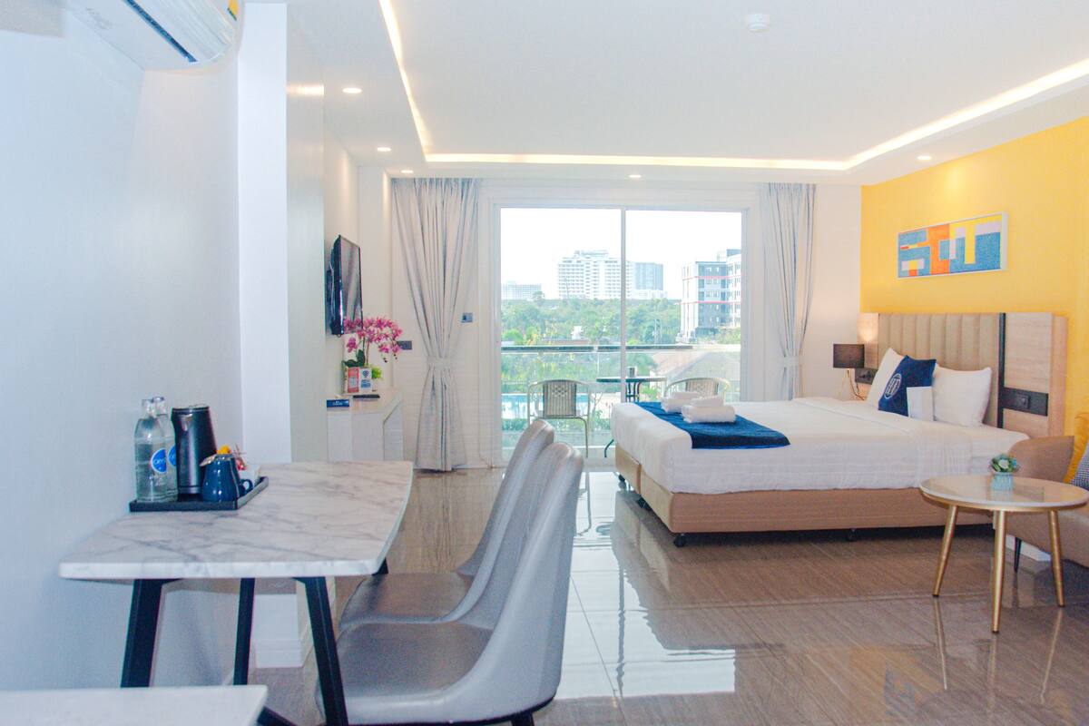 A hotel room with a bed, a desk, a chair, a small table, a TV, and a window with a view of buildings.