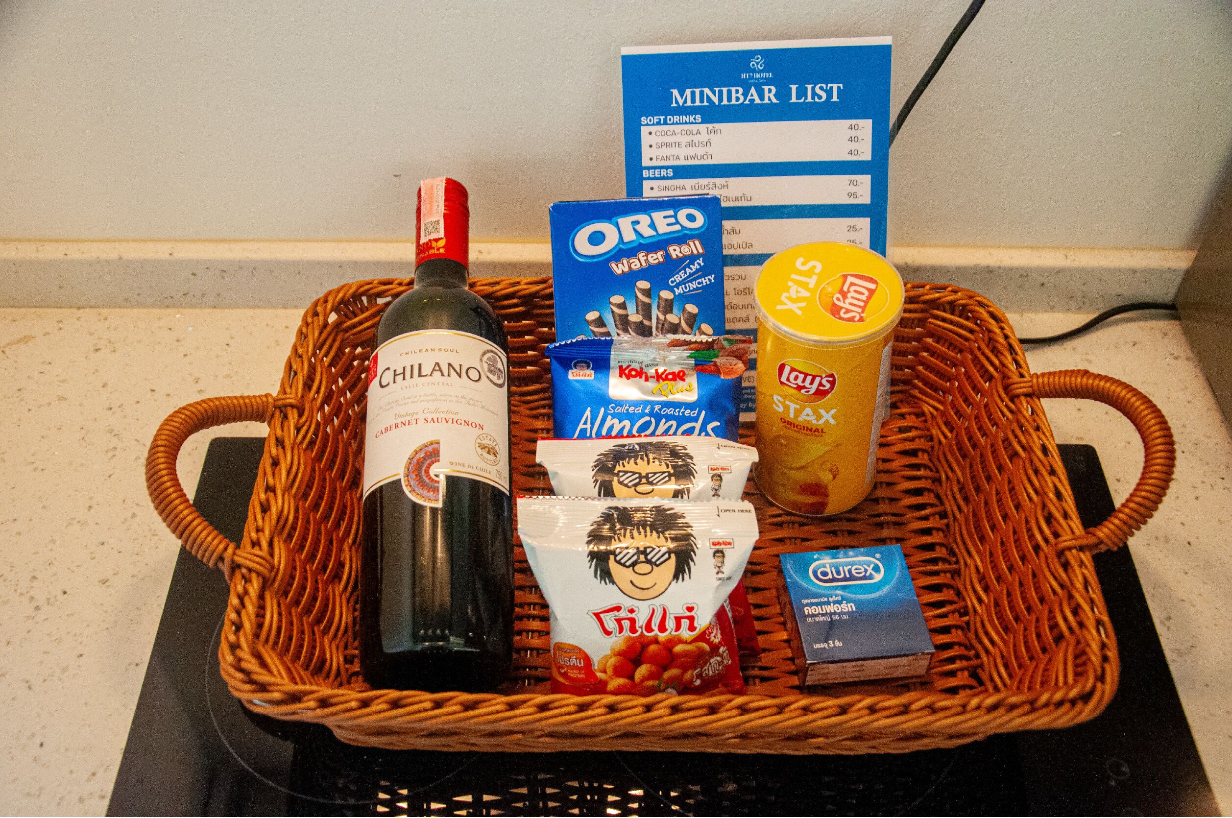 A wicker basket containing a bottle of Chianto wine, a box of Oreos, Lay's chips, a bag of chili, and a box of Durex condoms.