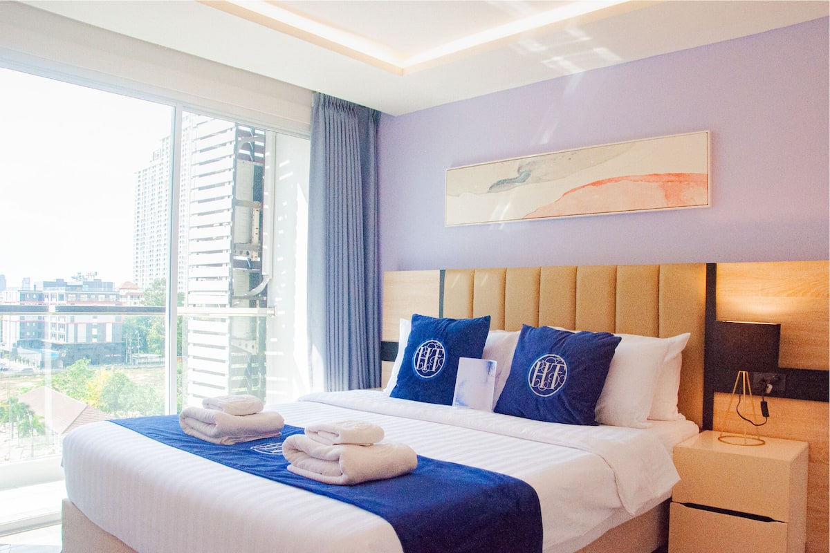 A hotel room with a large bed, a nightstand, a window with a view of buildings, and a painting on the wall.