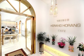 Interior de café com design moderno, apresentando entrada por porta de vidro, área de estar e plantas decorativas.