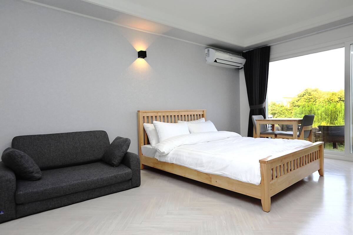 Ein modernes Schlafzimmer mit einem hölzernen Bett, einem grauen Sofa, einem kleinen Tisch und einem großen Fenster mit Blick auf Grünflächen.