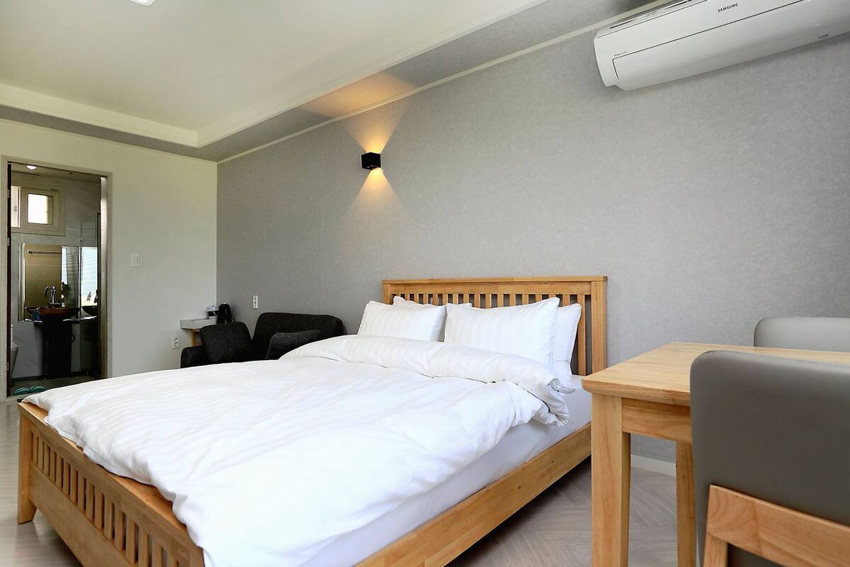 Ein ordentlich bezogenes Bett mit weißen Leinen, ein Holzkopfbrett und ein Nachttisch mit Stuhl. Eine Klimaanlage ist an der Wand über dem Bett angebracht.