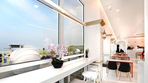 A modern hotel interior with a dining area, a large window with a view of the beach, and a potted orchid on the windowsill.