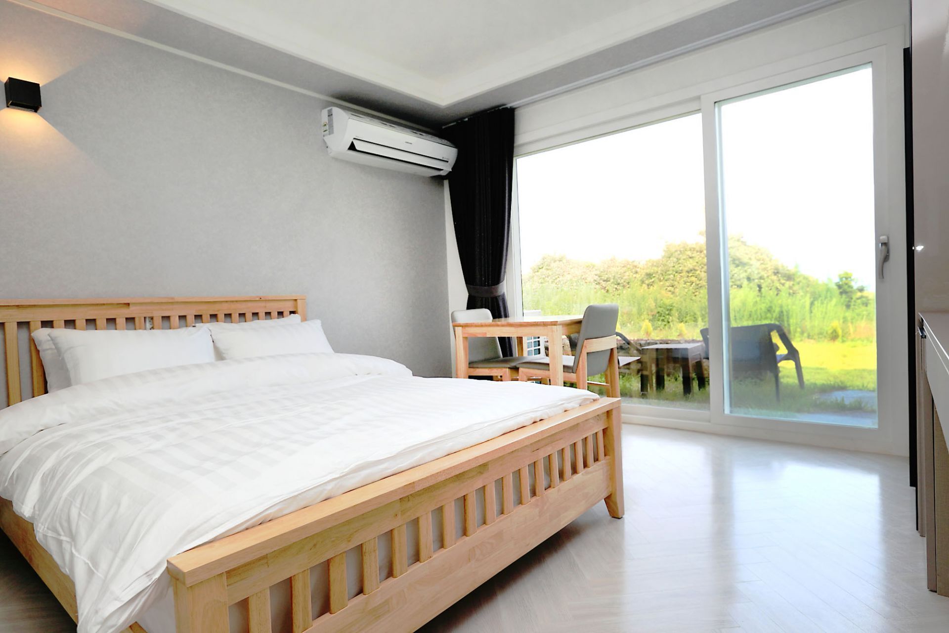 A bedroom with a wooden bed, a desk, and a chair, and a sliding glass door leading to a balcony with a view of greenery.