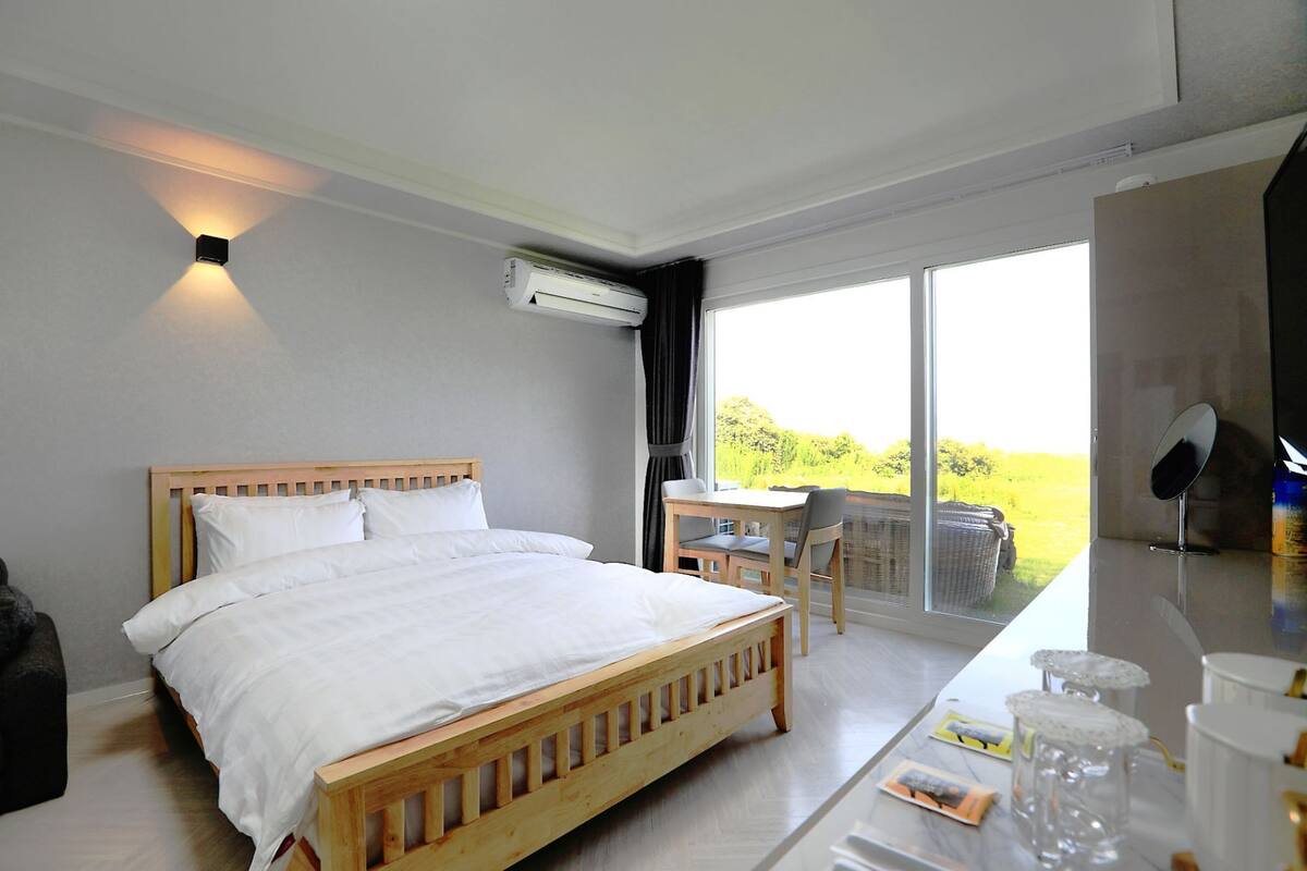 Ein Schlafzimmer mit einem hölzernen Bett, einem kleinen Tisch mit Stühlen, einem großen Fenster mit Blick auf Grünflächen und einer Klimaanlage an der Wand.