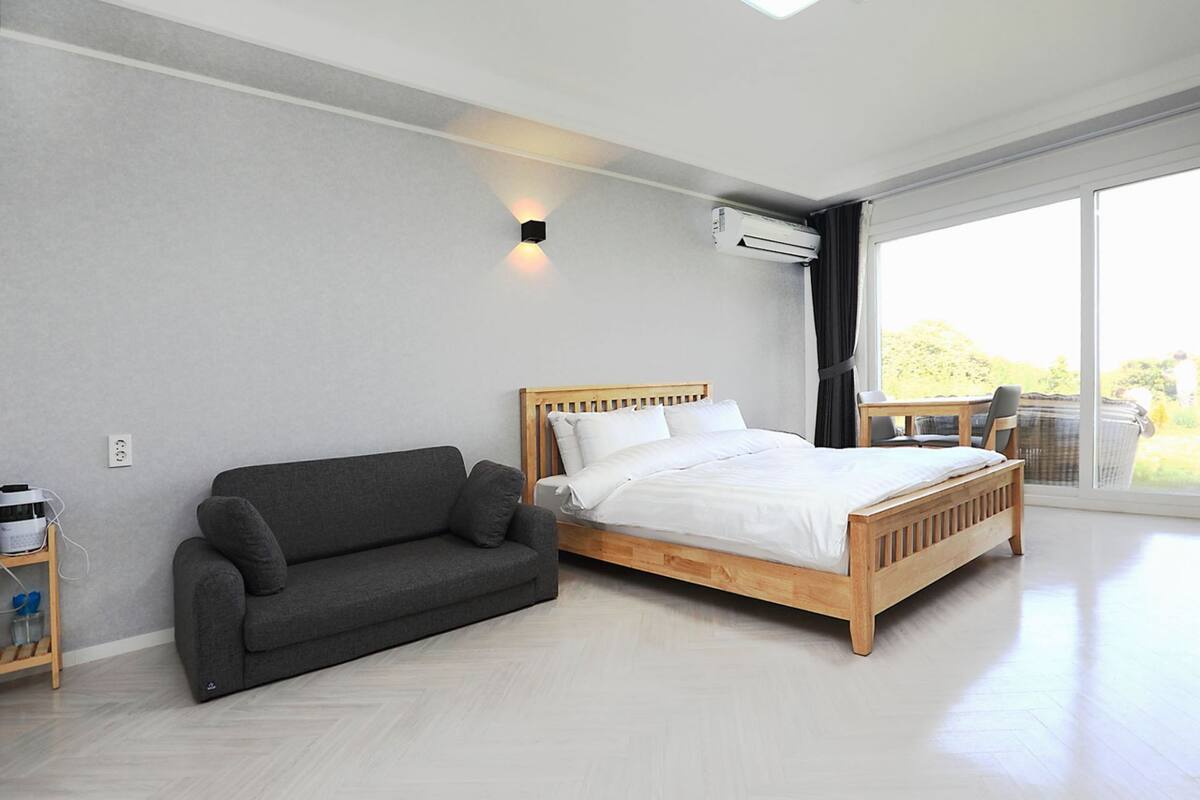 Ein modernes Schlafzimmer mit einem hölzernen Bett, einem grauen Sofa und einem großen Fenster mit Blick ins Freie.
