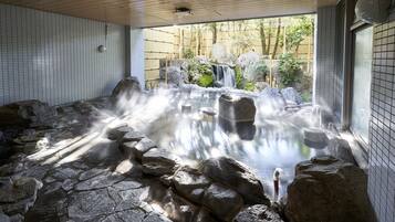 A hot spring bath area with stone flooring, a waterfall feature, and a view of the outdoors through a window.