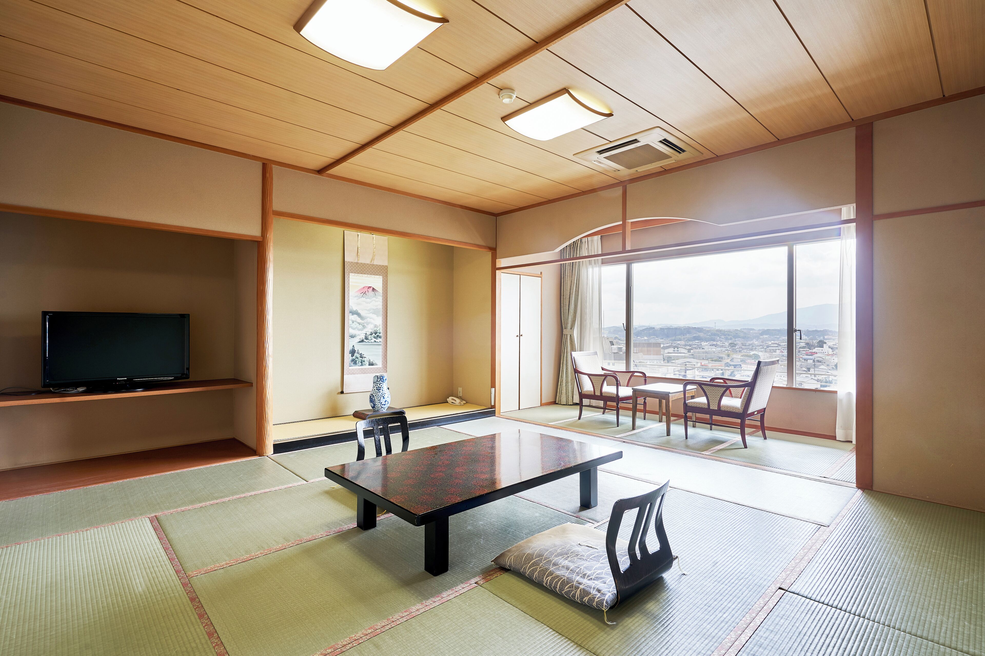 Una habitación japonesa tradicional con suelo de tatami, una mesa baja, sillas, un televisor y un ventanal con vistas a la ciudad.
