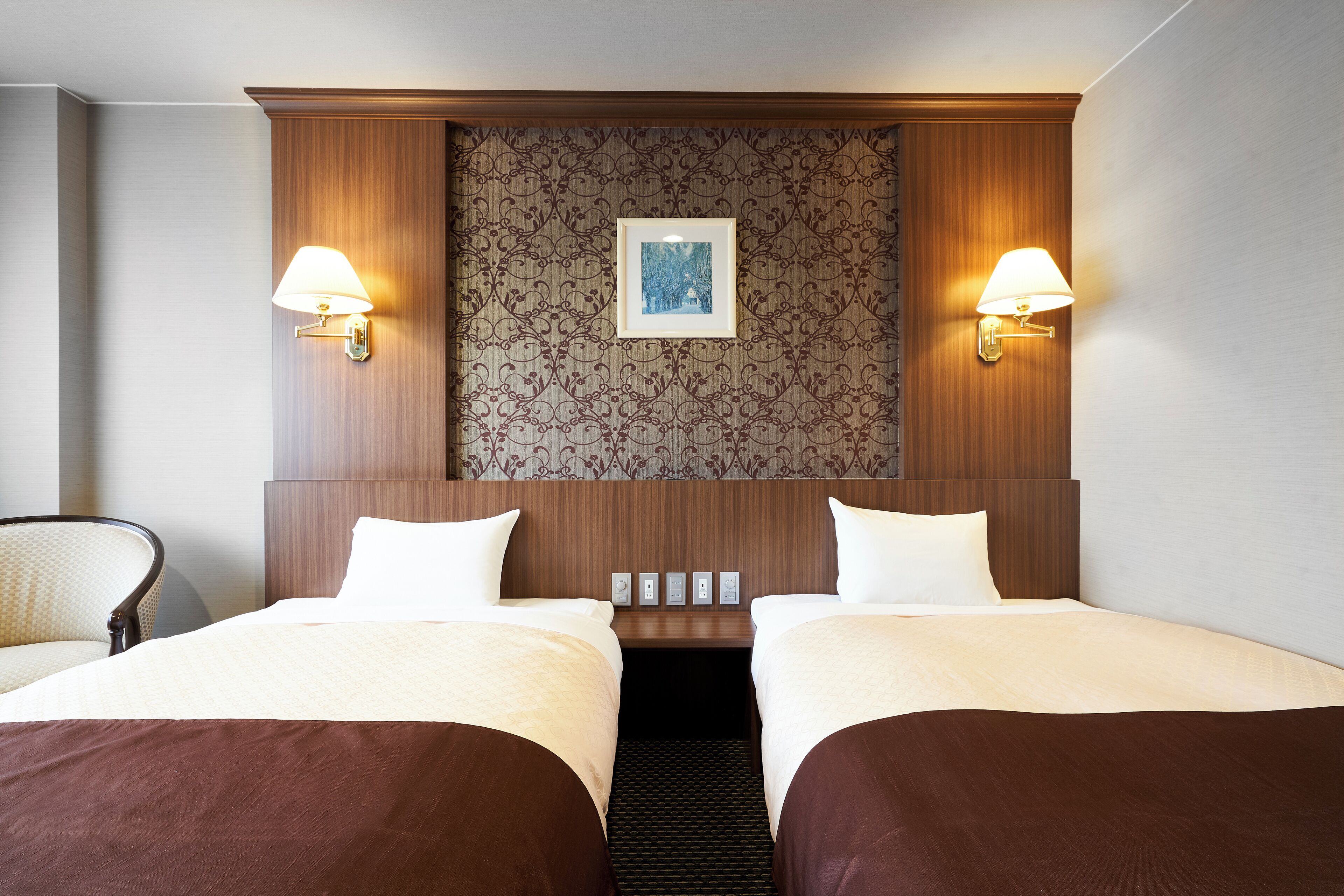 Habitación de hotel con dos camas, cabecera con estampado floral, lámparas fijadas a la pared y un cuadro enmarcado.