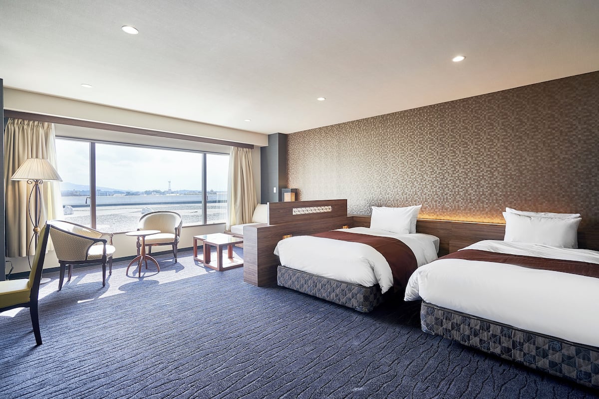Een hotelkamer met twee bedden, een bureau, een stoel en een groot raam met uitzicht op de stad.