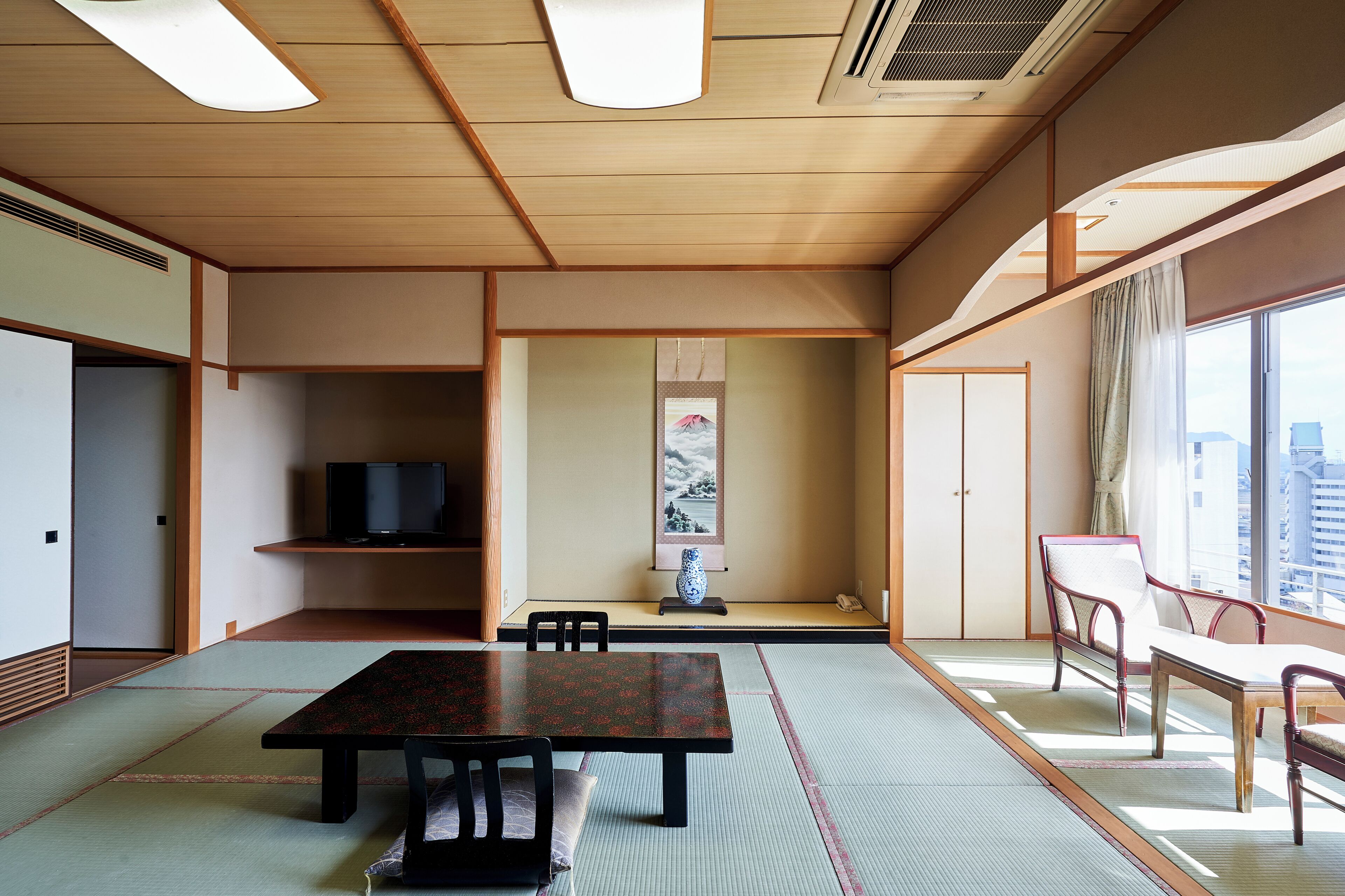 A traditional Japanese room with tatami flooring, a low table, chairs, a television, and a view of the city.