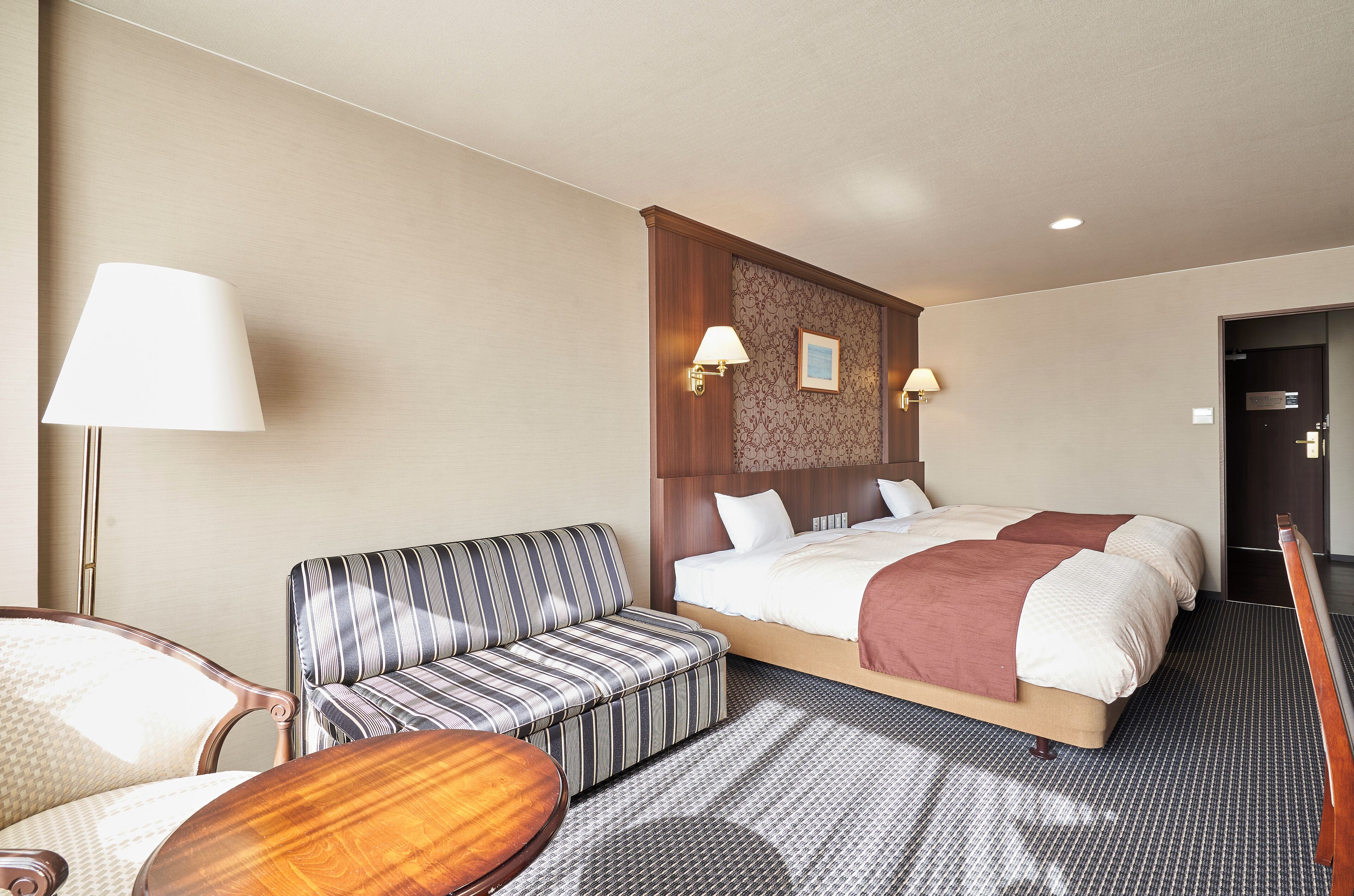 A hotel room with a bed, a striped sofa, a wooden side table, and two wall-mounted lamps.