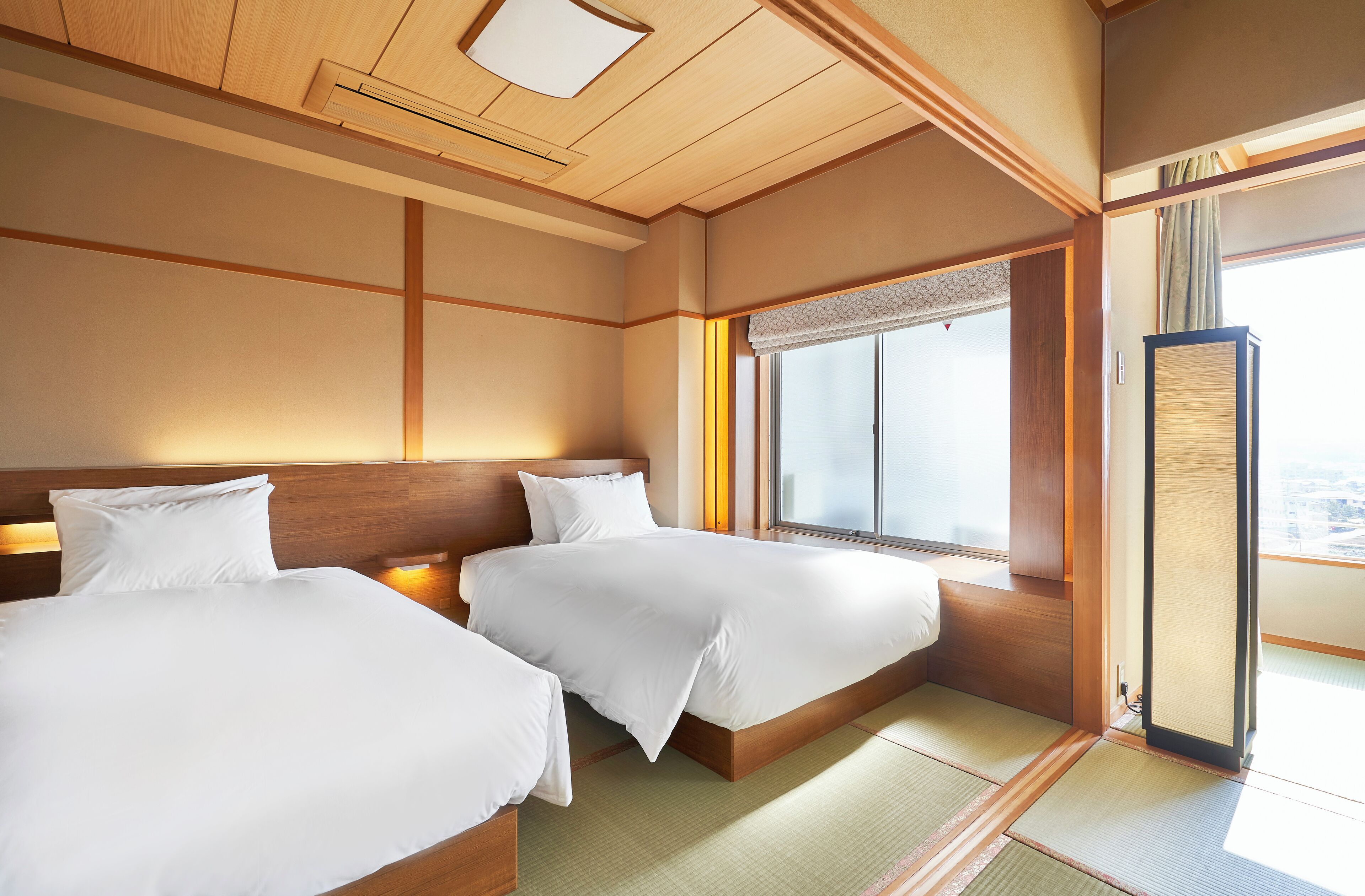 Une chambre d’hôtel avec deux lits, des têtes de lit en bois, une grande fenêtre avec vue, un sol recouvert de tatamis et une porte coulissante de style japonais traditionnel.