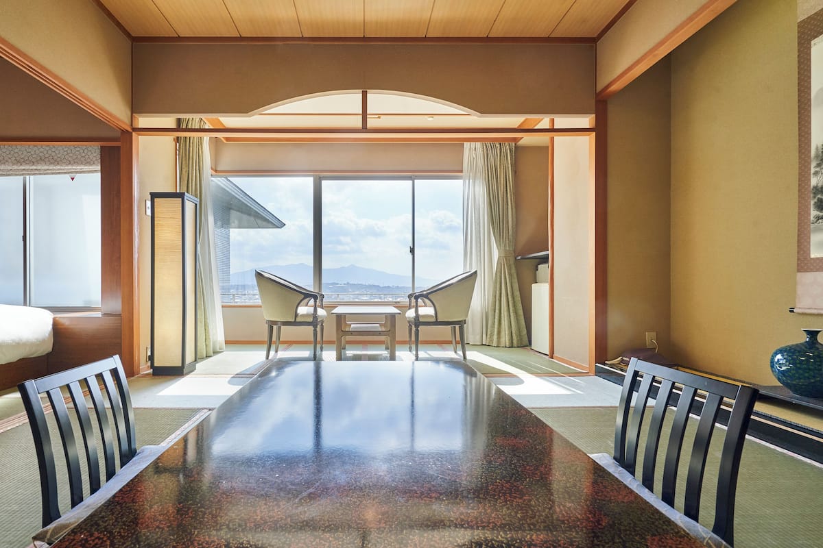 Een hotelkamer met een grote eettafel, twee stoelen, een bed en uitzicht op de stad door grote ramen.