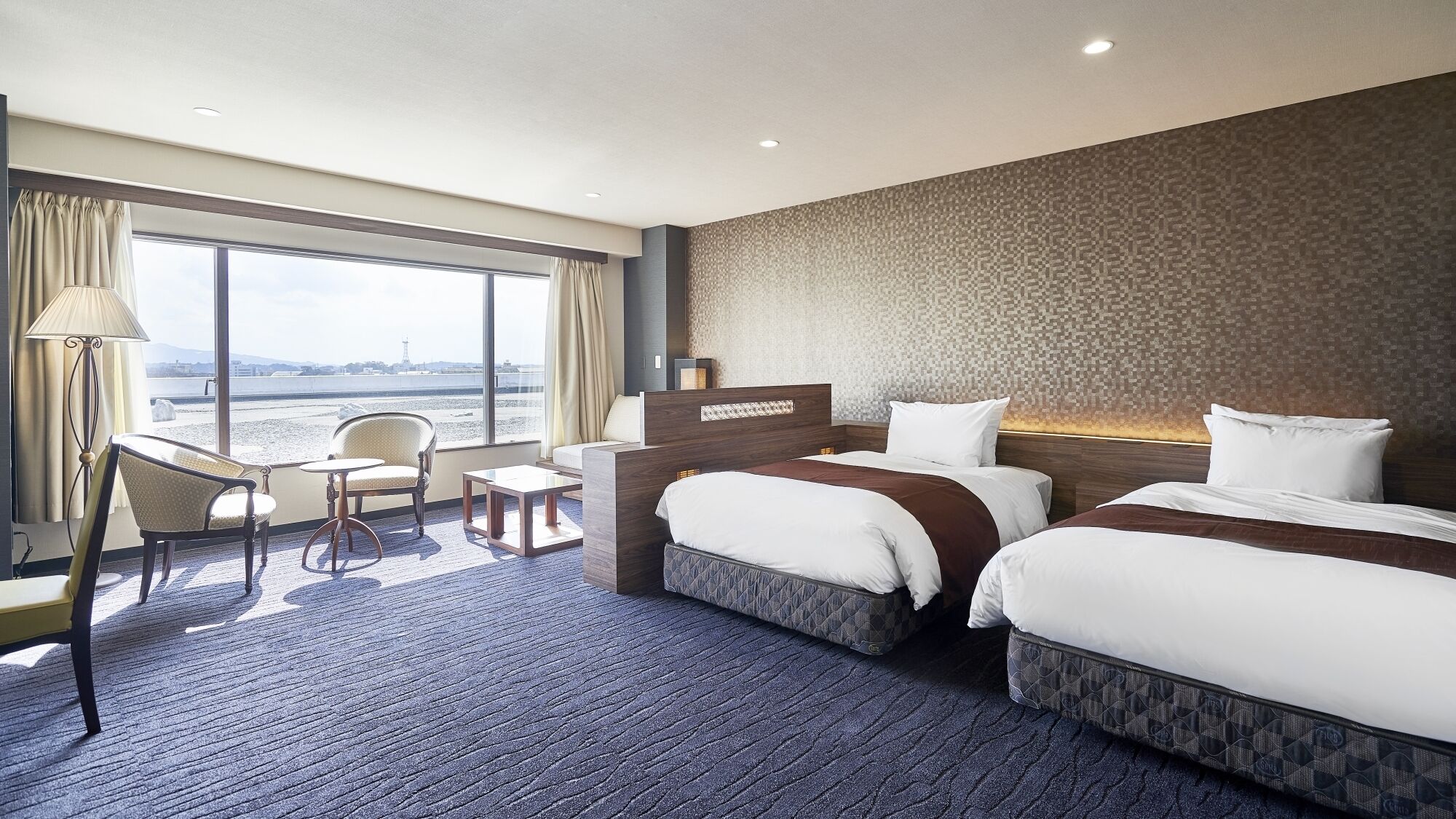 Une chambre d’hôtel avec deux lits, un bureau, une chaise et une grande fenêtre donnant sur un beau paysage.