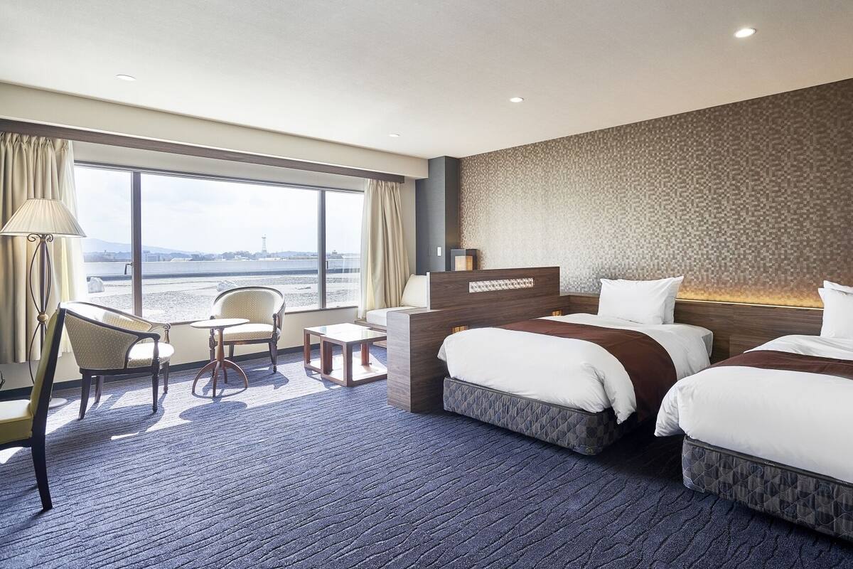 Een hotelkamer met twee bedden, een bureau, een stoel en een groot raam met uitzicht.