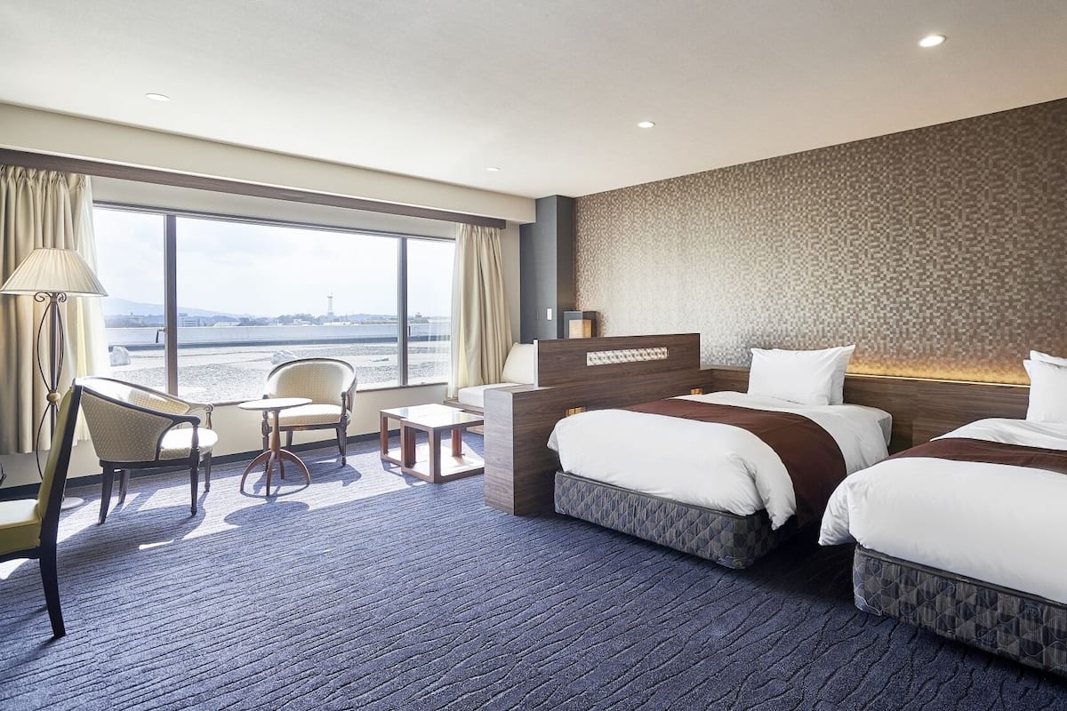 Habitación de hotel con dos camas, un escritorio, una silla y un ventanal con vistas.