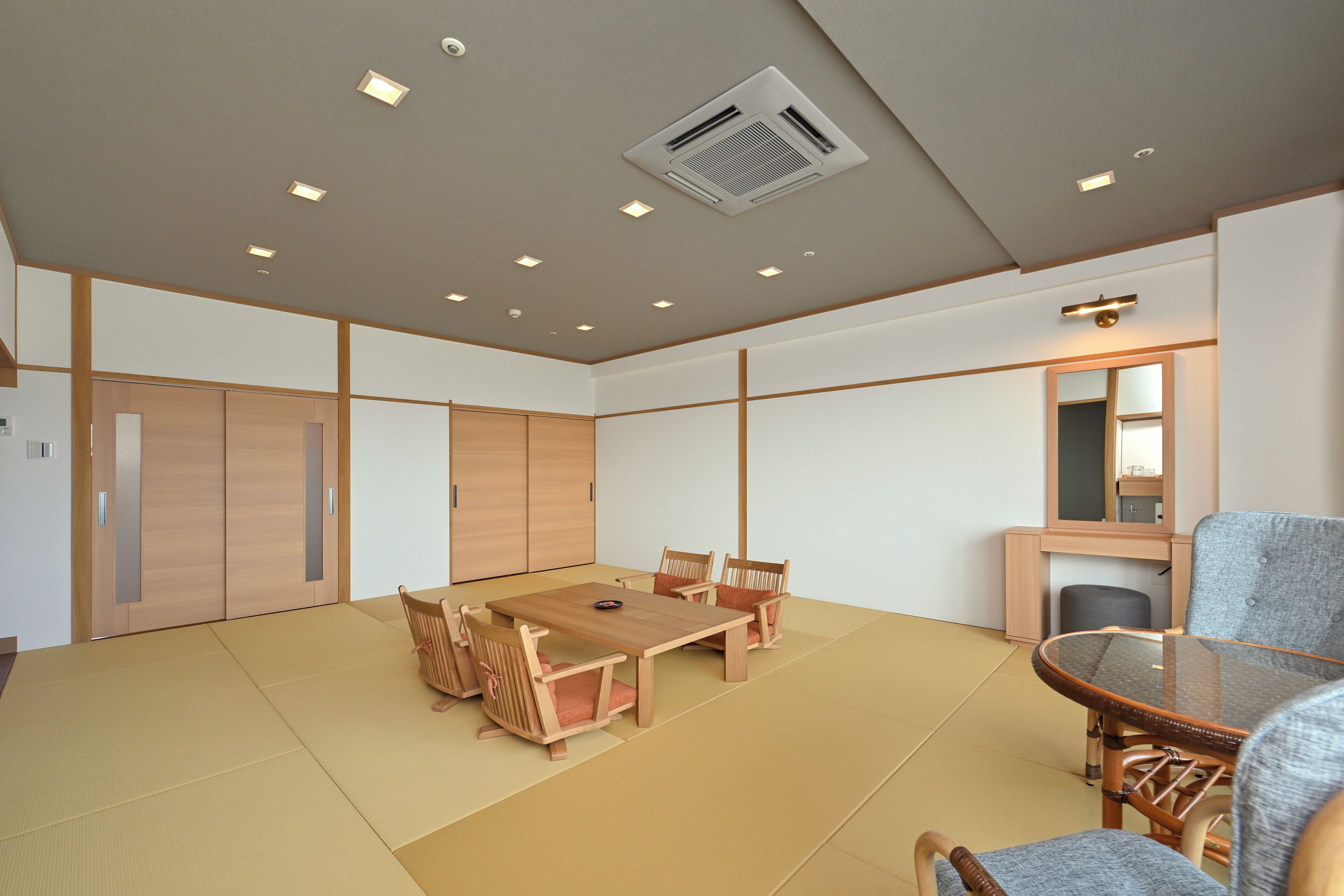 Una stanza con pavimento in tatami, un tavolo da pranzo in legno con sedie, un piccolo tavolino rotondo con una sedia, uno specchio e una bocchetta di ventilazione sul soffitto.